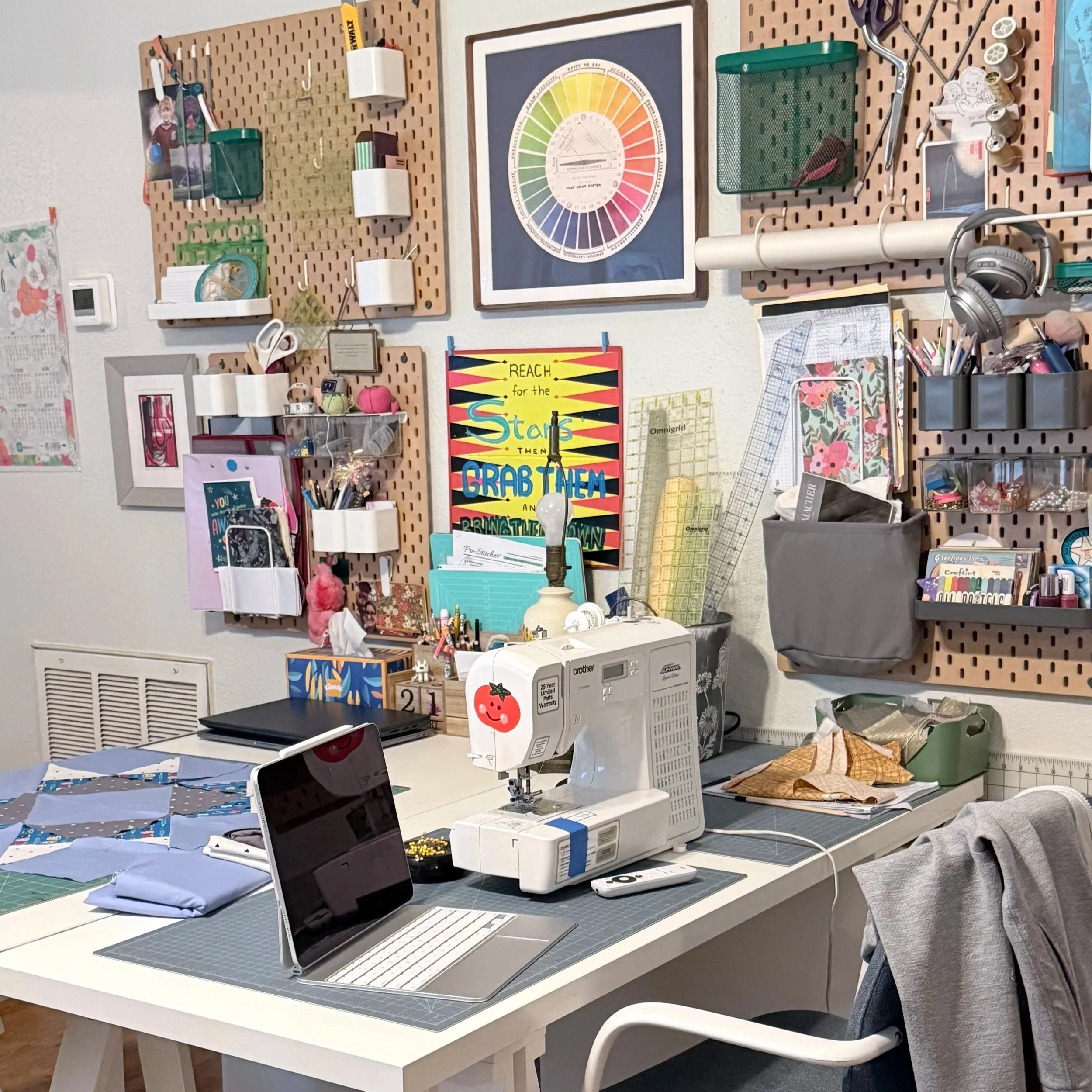 A crafting workspace with a white table, computer, fabric, and supplies. Wooden pegboard wall with tools, scissors, thread, and inspirational posters.