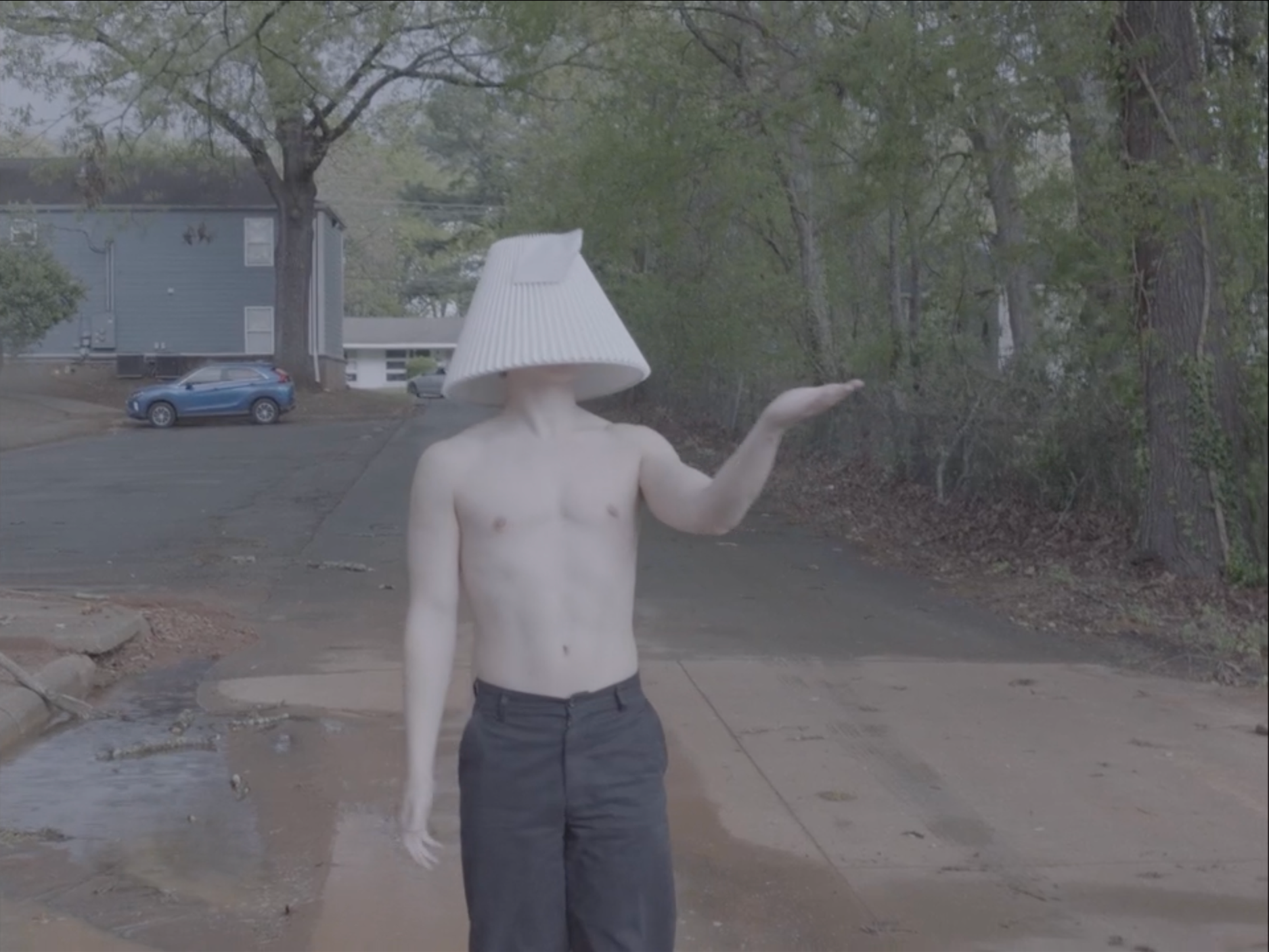 A still from the film "Lamp Man." A person with no shirt, wearing dark pants, has a lamp shade over their head and is standing in a driveway with trees and houses in the background.