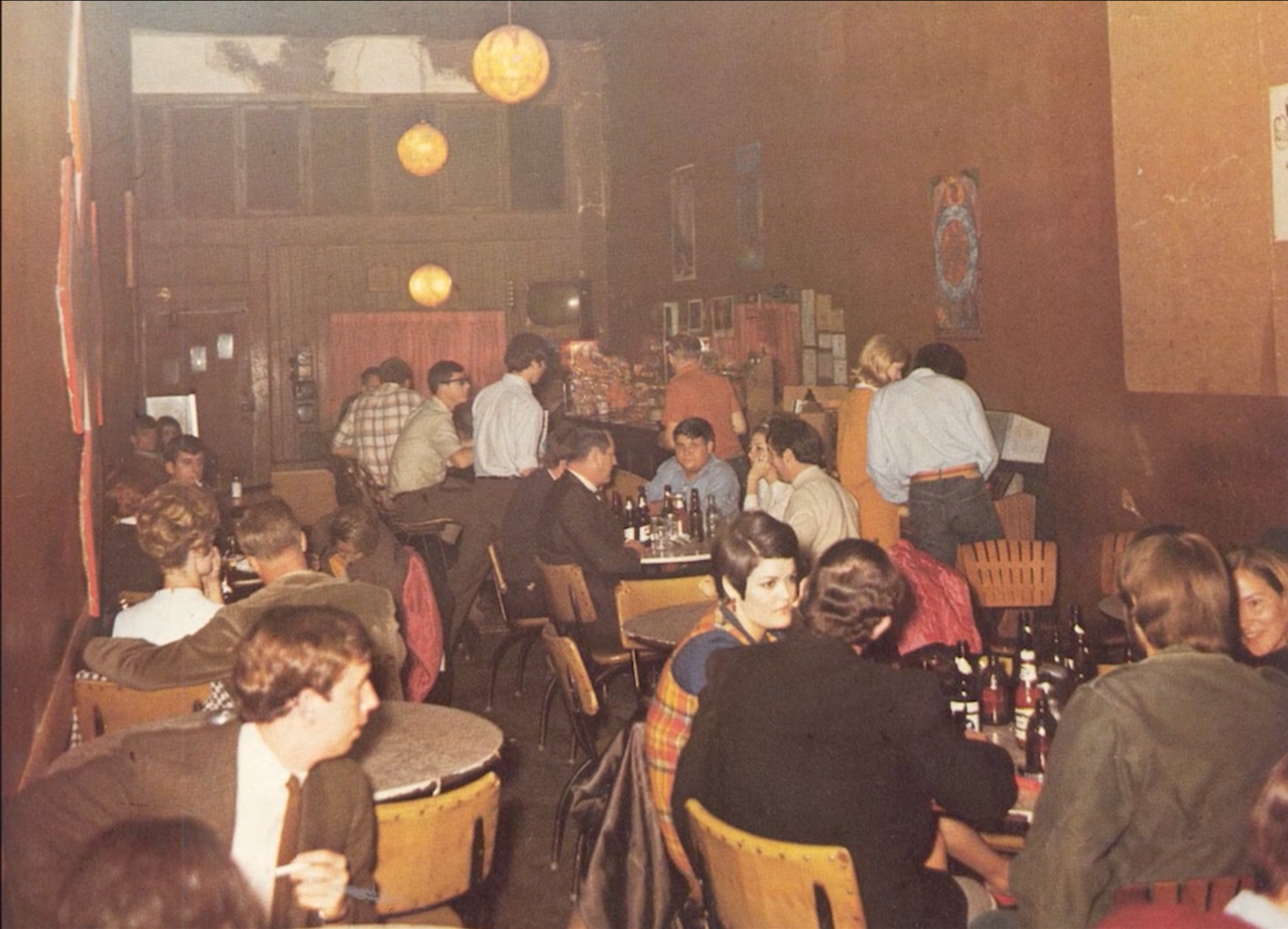 A still from the film "The Chukker." Vintage photo of a crowded bar with people socializing, sitting at tables, and standing in line, decorated with warm lighting and artwork on the walls.