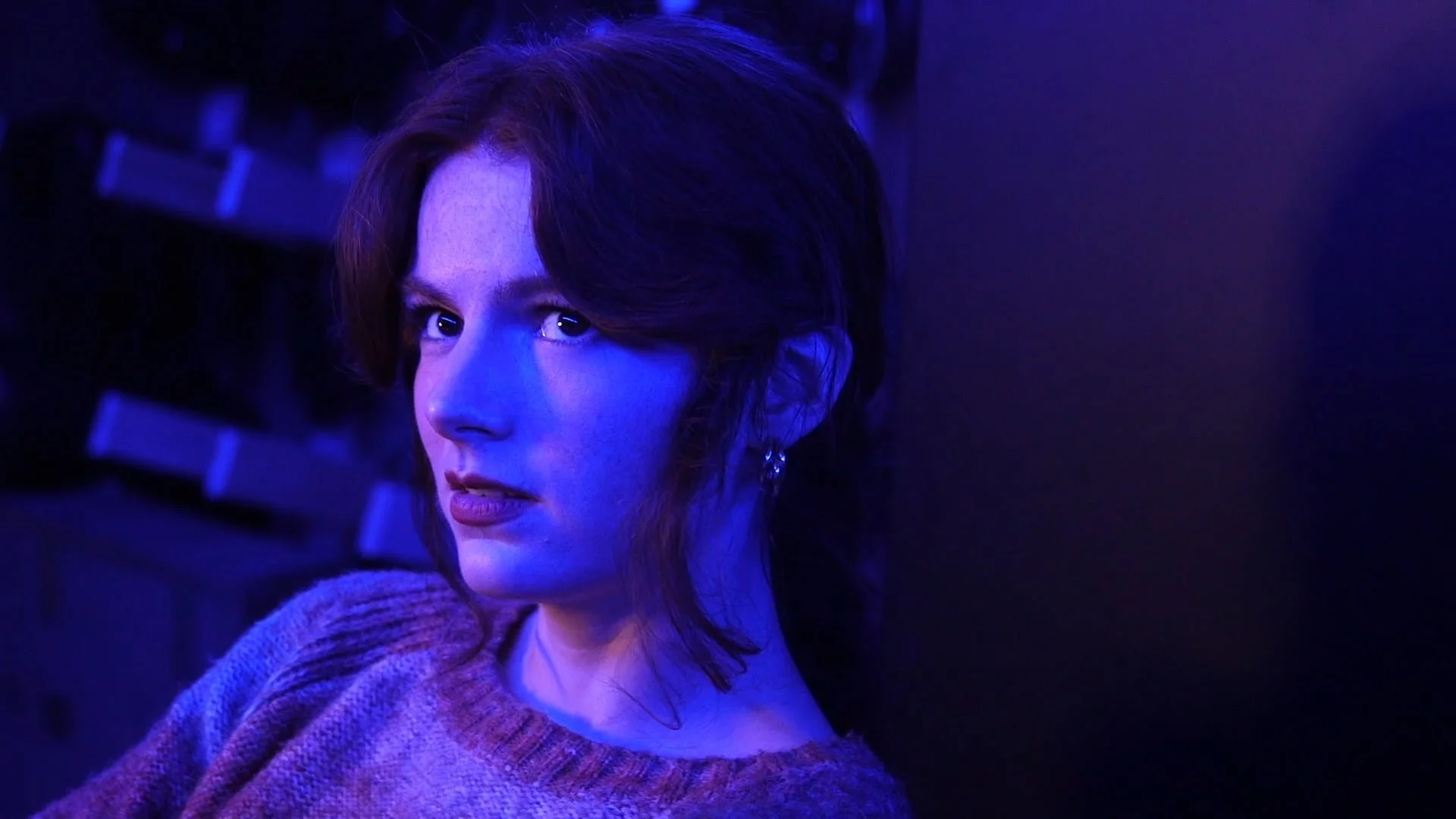 A still from the film "The Undiscovered Country." A woman with short brown hair and earrings poses against a dark background illuminated by blue and purple lighting.