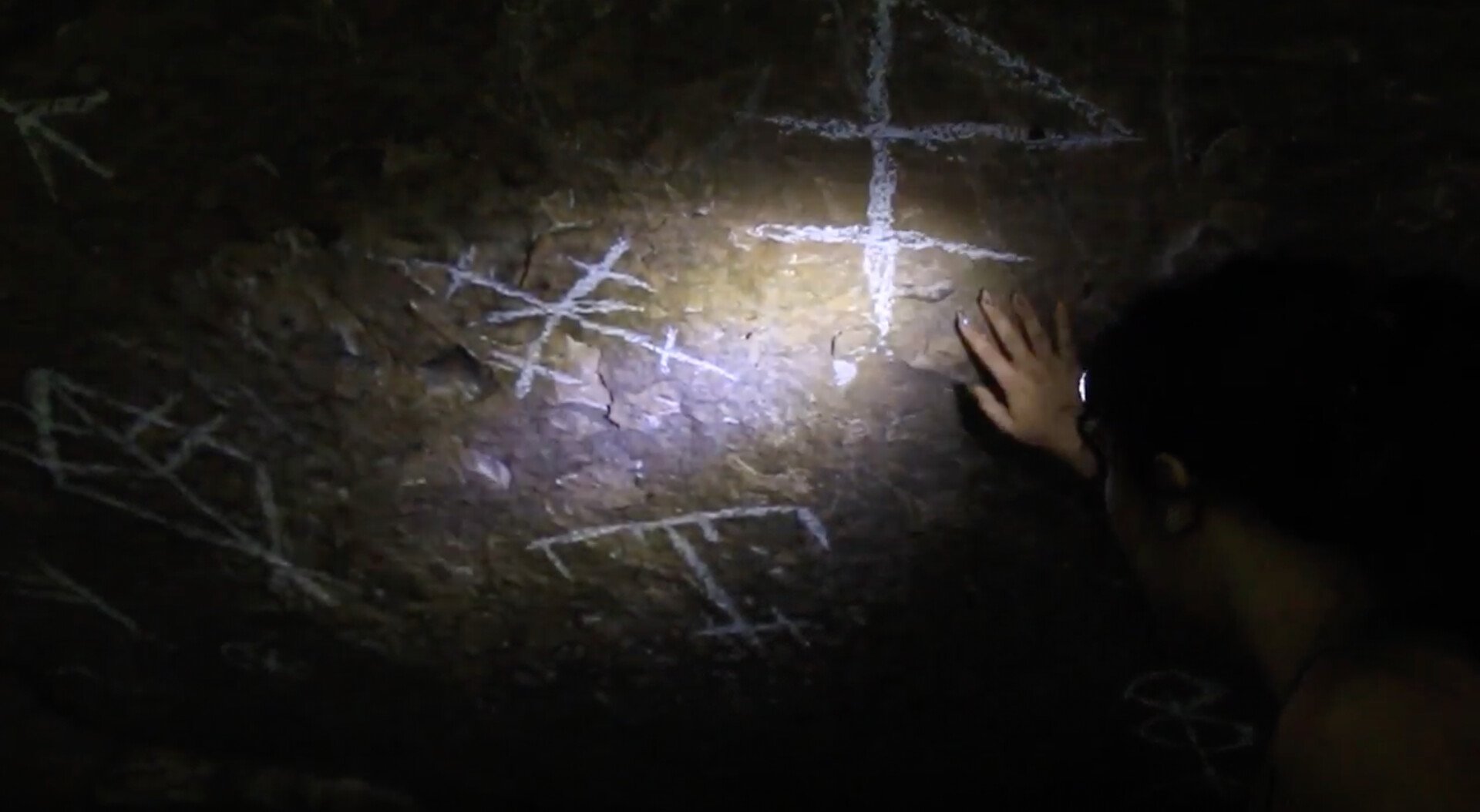 A still from the film "Echoes of the Unknown." Person exploring a dark cave, marking scope with white chalk, hand visible near the markings.