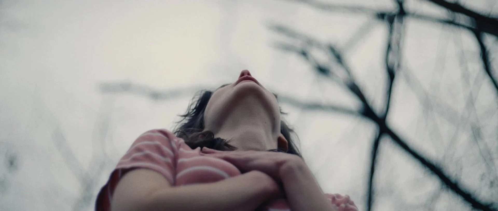 A still from the film "Cliff."  A woman with dark hair wearing a pink striped shirt looking up with her hand on her chest, with a background of blurred leafless trees and an overcast sky.
