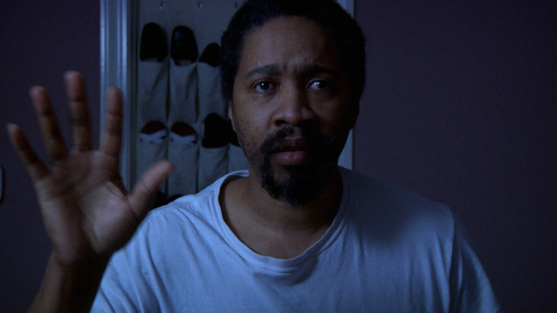 A still from the film "What Stares Back." A man with dark hair and a beard raises his right hand as if to stop or say hello.