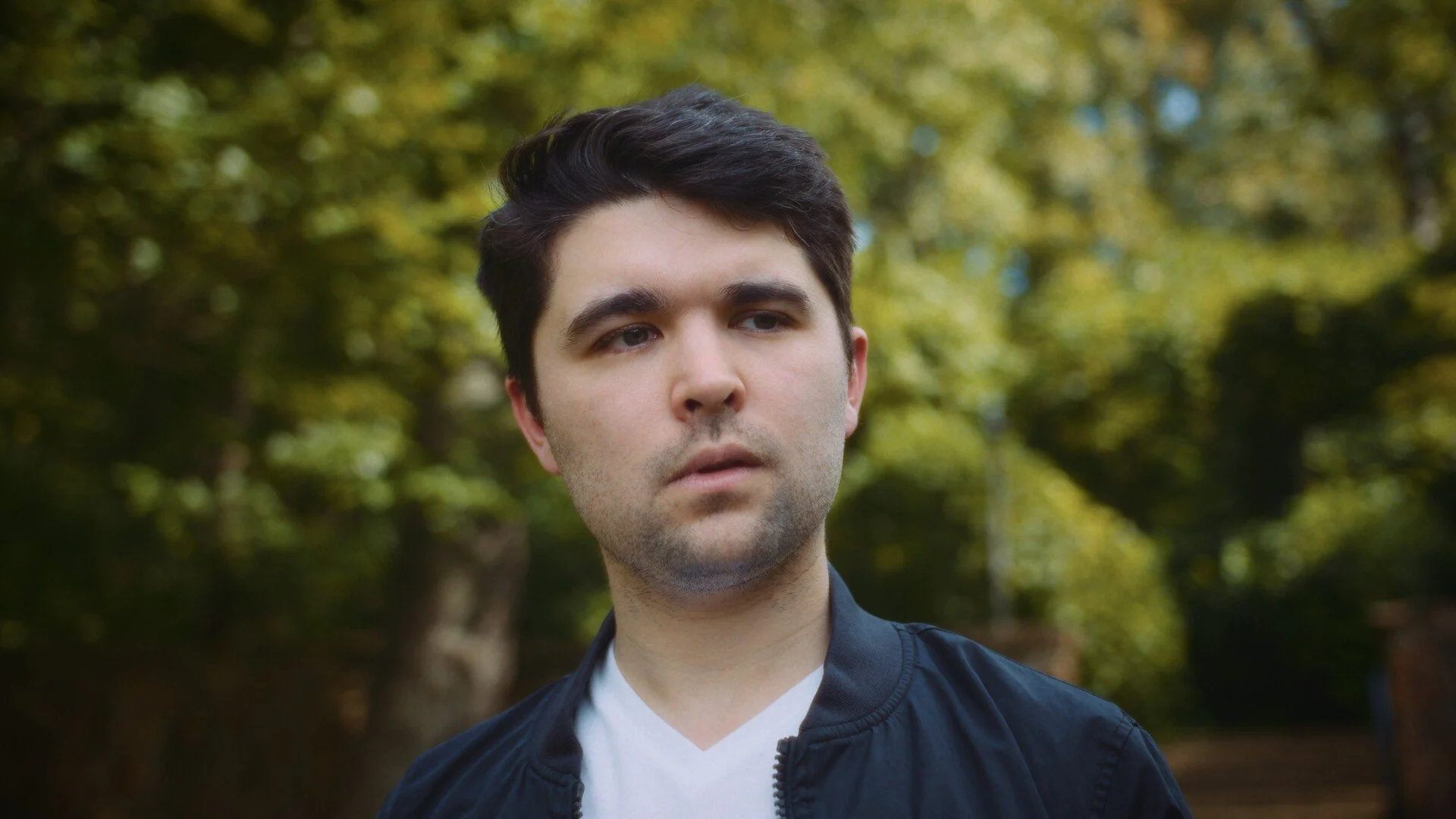 A still from the film "Mel." A young man with dark hair and light skin is outdoors in front of trees with green and yellow leaves.