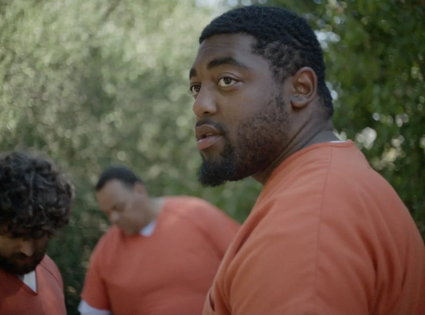 A still from the film "All the Real Boys." A man with a beard and short curly hair looking to the side, wearing orange prison attire.