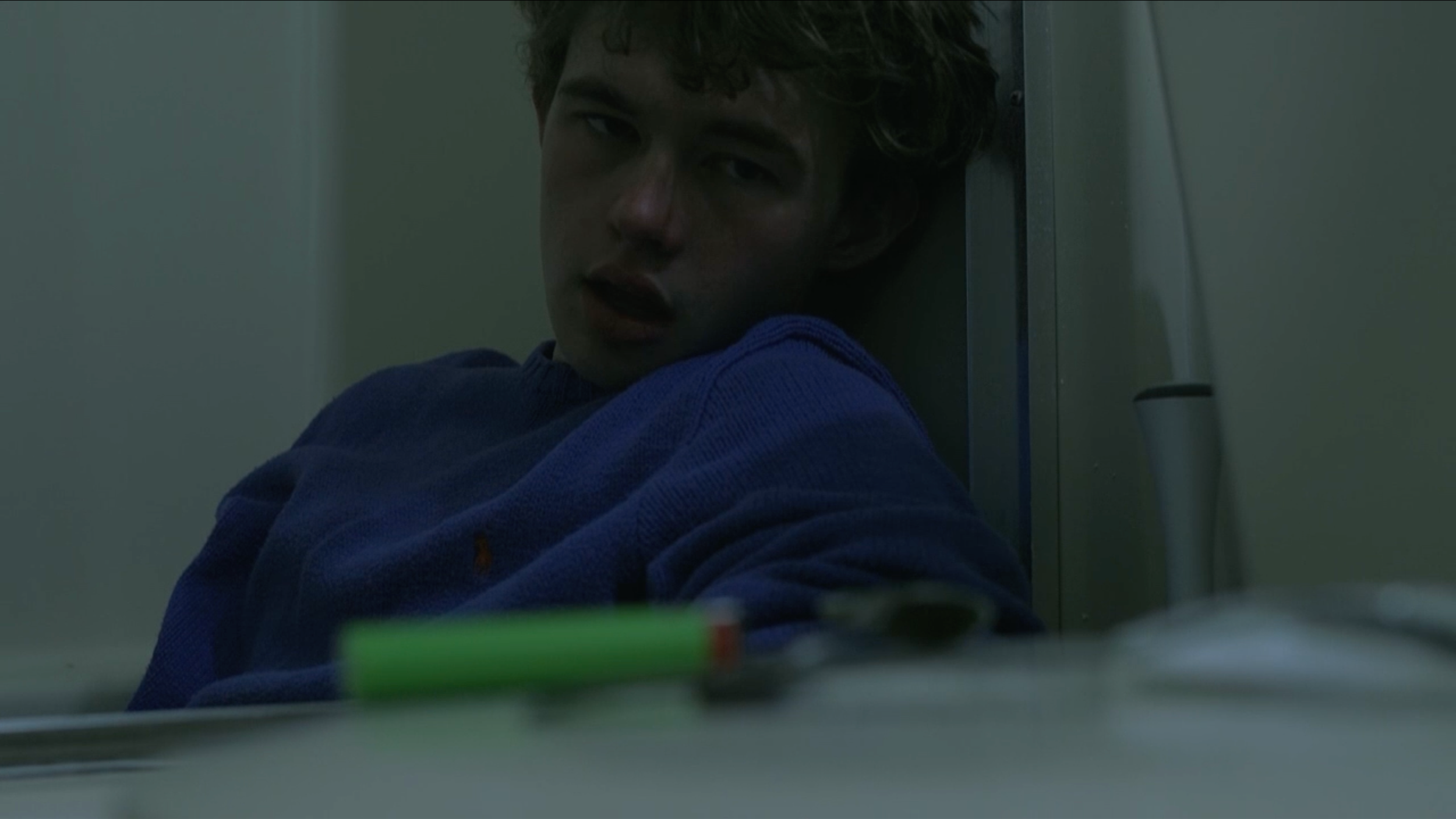 A still from the film "Exit." A young man with curly hair and a blue sweater, sitting against a wall with a distressed expression, looking at the camera.