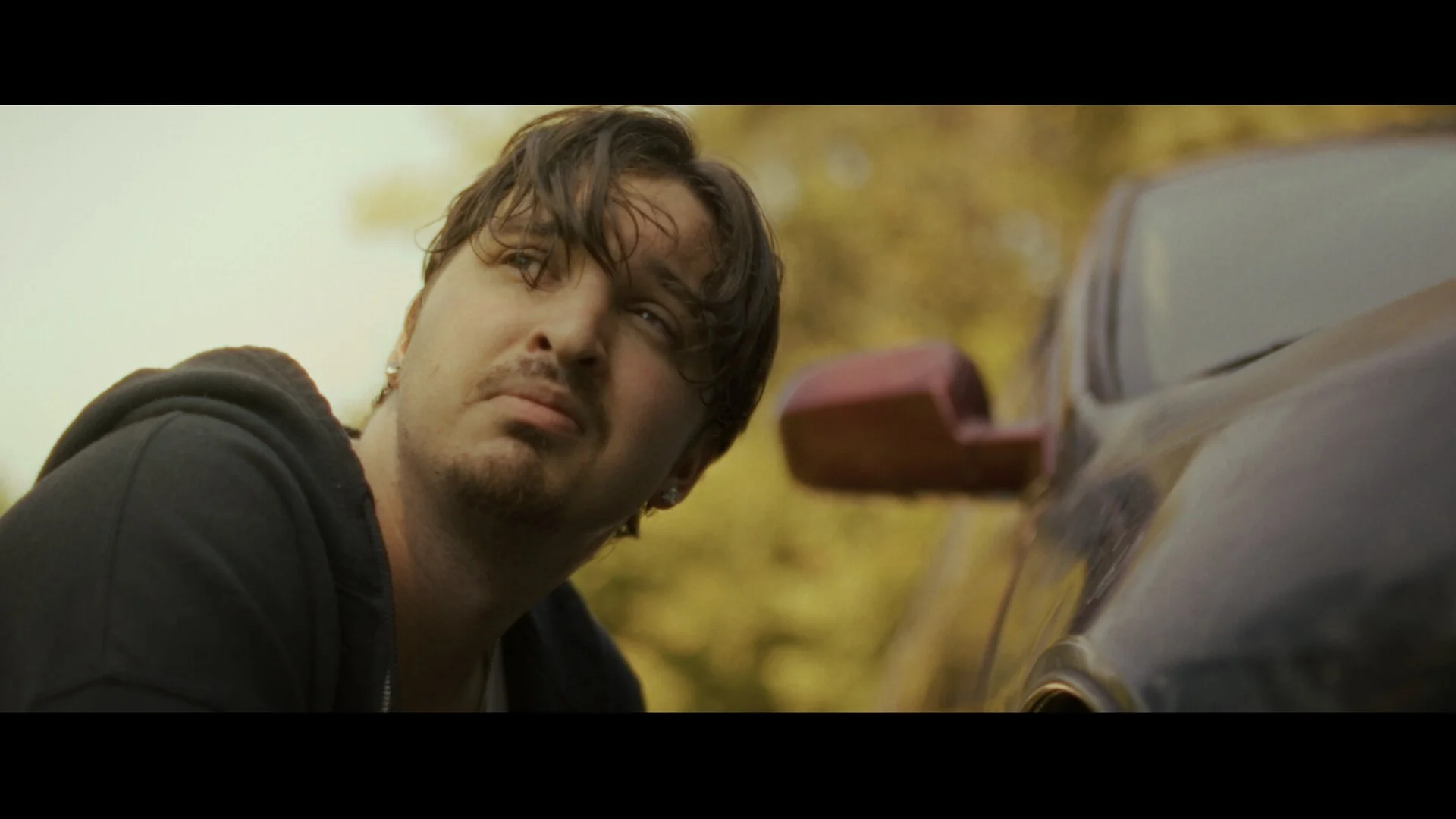 A still from the film "Somewhere Off 72." A man with dark hair and slight stubble looking to the side outdoors near a black car with a red side mirror; background features yellow foliage.