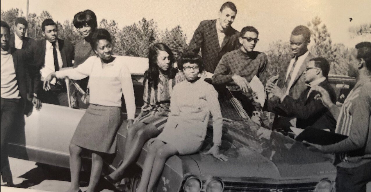 A still from the film "The First to Walk In." A black-and-white photo of a group of people gathered around a car.