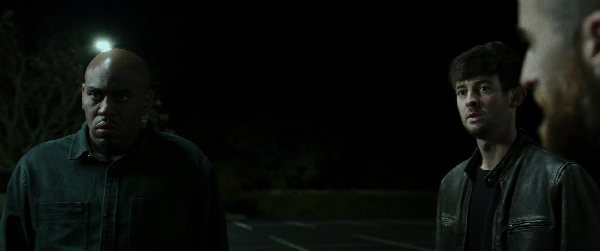 A still from the film "Closer Than A Brother." Three men standing outdoors at night, appearing serious or concerned, with trees and a parking lot in the background.
