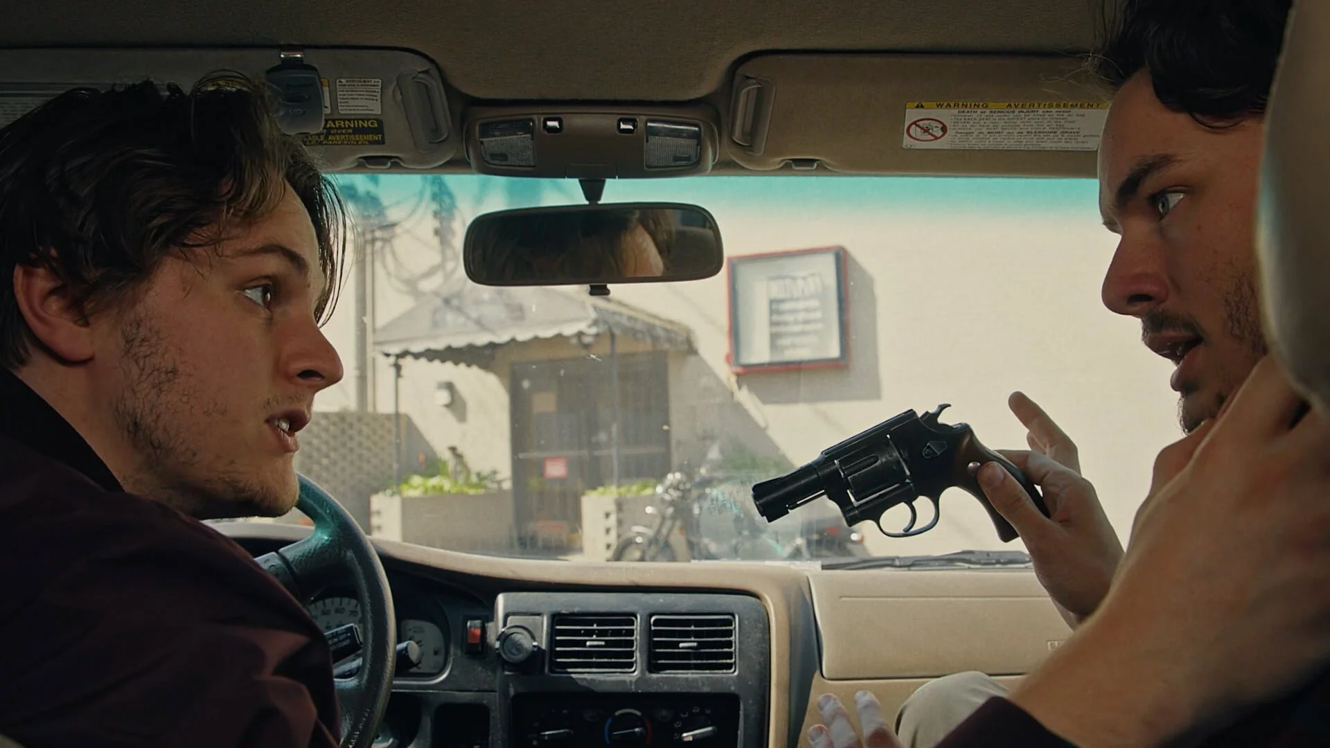 A still from the film "Robbers." Two men inside a vehicle, one holding a gun and pointing it at the other, who appears surprised.