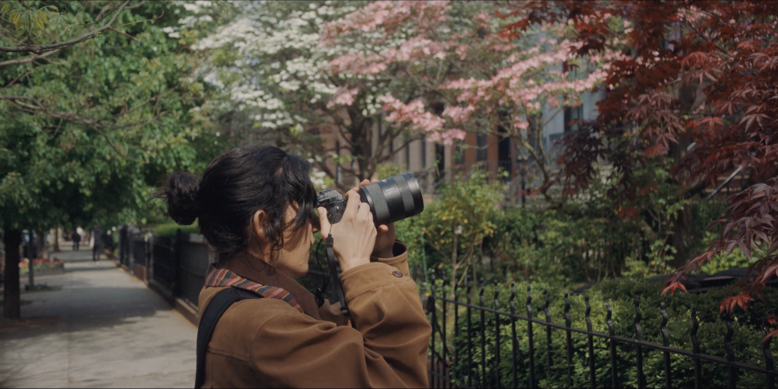 A still from the film "The Scout." A woman with dark hair takes photos with a camera on a sidewalk.