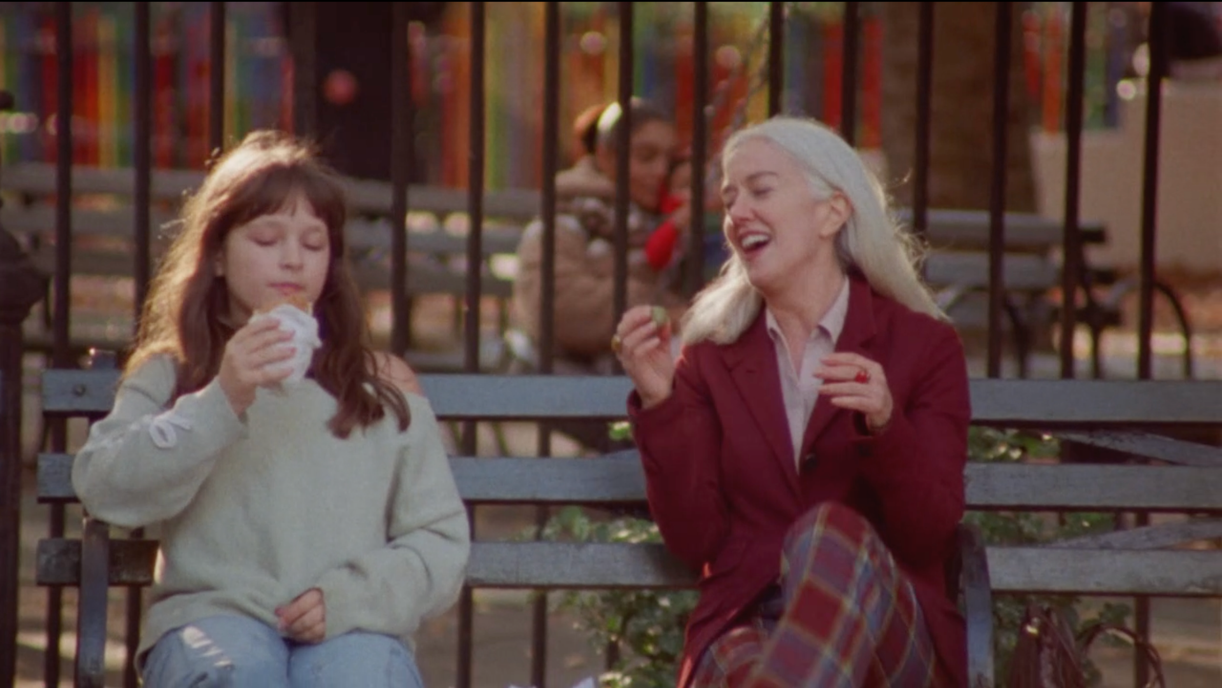 A still from the film "Olive Juice." A young girl and an older woman sitting on a park bench, with children playing in the background.