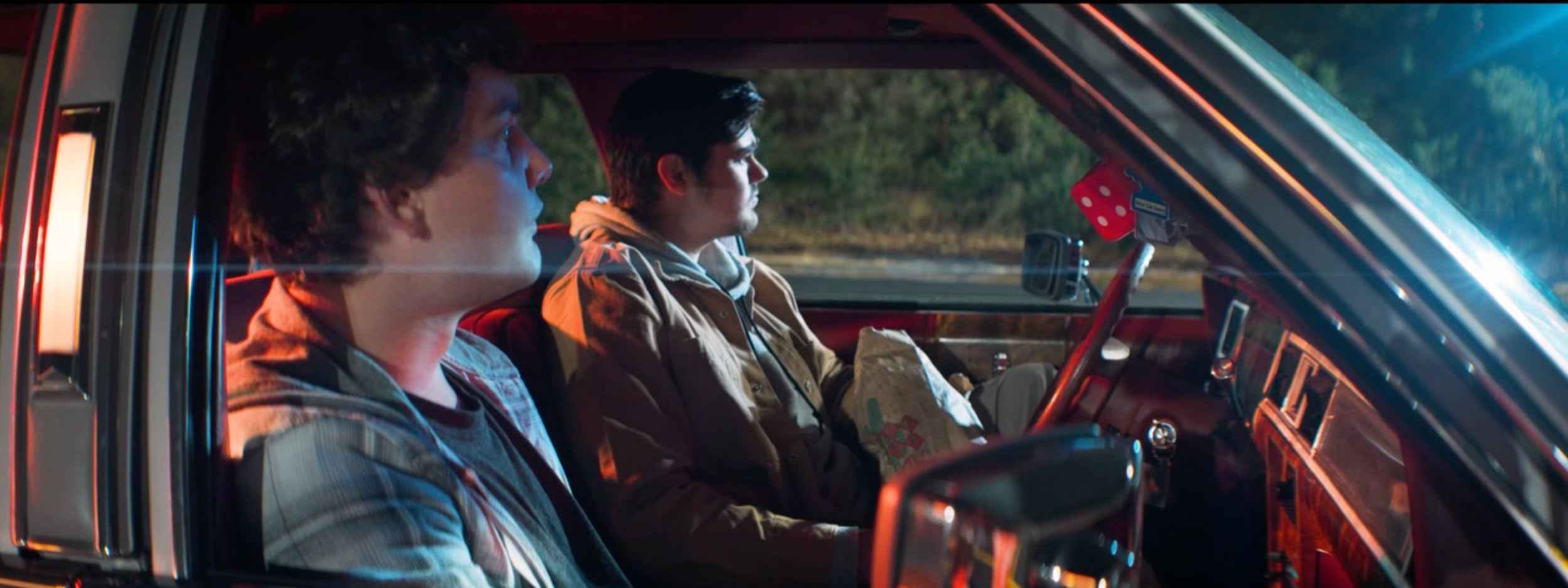 A still from the film "Return to Sender." Two young men sitting inside a car at night, illuminated by colorful interior and exterior lights.
