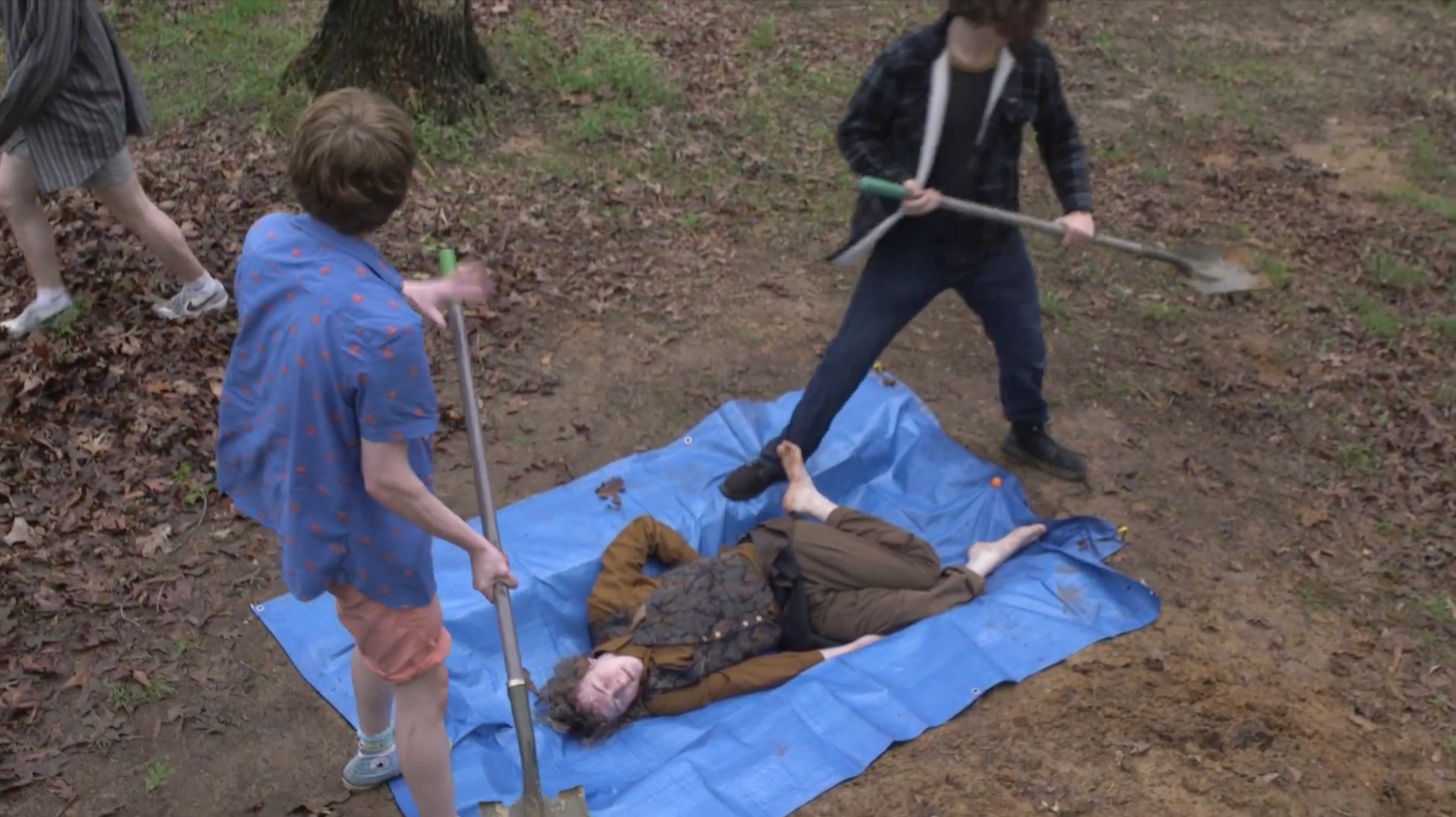 A still from the film "The Mysterious Death of Gobblestone Stone Gobble." Multiple people surrounding a person lying on a blue tarp in a wooded area, one person raising a shovel.