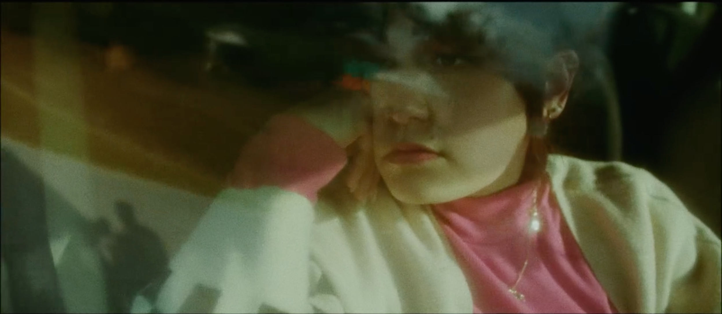 A still from the film "Raya." A woman looking out of a car window, partially obscured by the reflection and a blurry windshield.