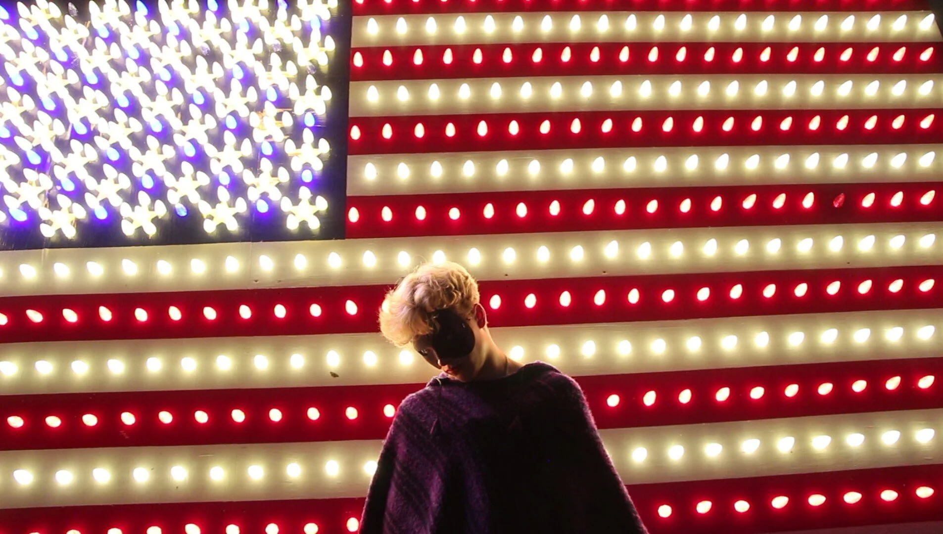 A still from the film "Return of the Purple Menace." A person wearing a mask stands in front of a large illuminated American flag sign, with red, white, and blue lights arranged in stripes and stars.