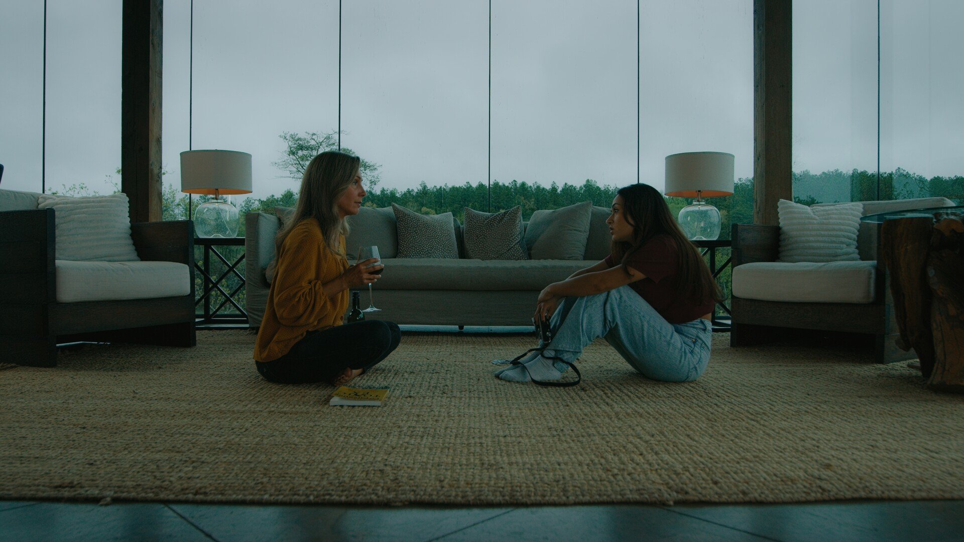 A still from the film "Little Eye." Two women sitting on the floor facing each other in a living room with large glass windows. One woman is holding a glass of wine, and the other has a camera.