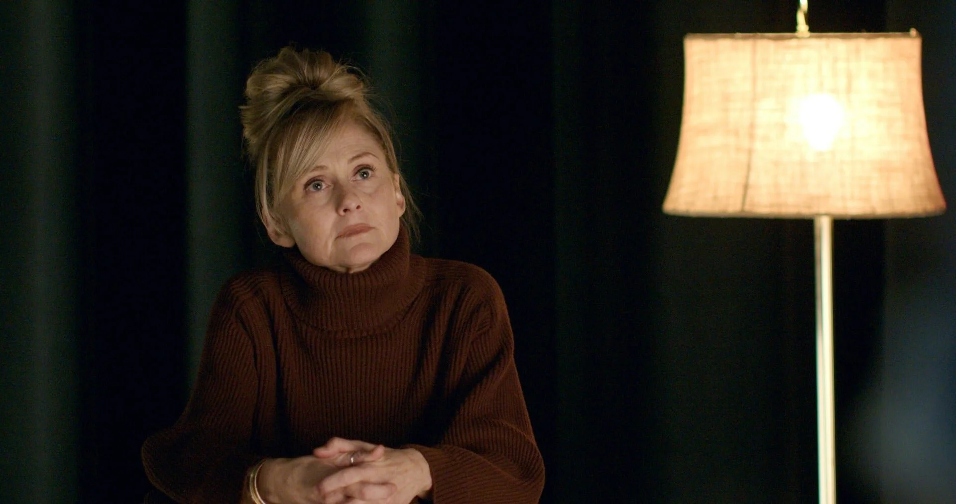 A still from the film "Leads." A woman with blonde hair styled in an updo, sitting with her hands clasped, looking thoughtful. A lamp with a beige shade is lit in the background against a dark backdrop.