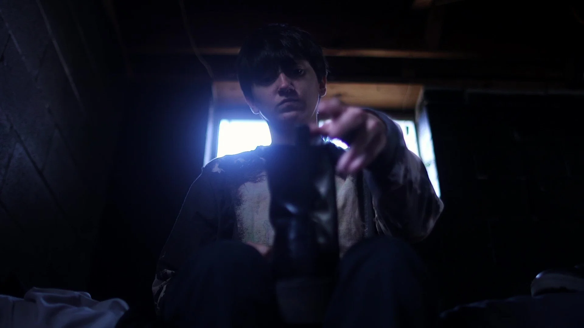 A still from the film "there's something watching me, but i don't know his name." A young person with dark hair pointing directly at the camera, sitting in a dimly lit room with a window glowing with natural light.