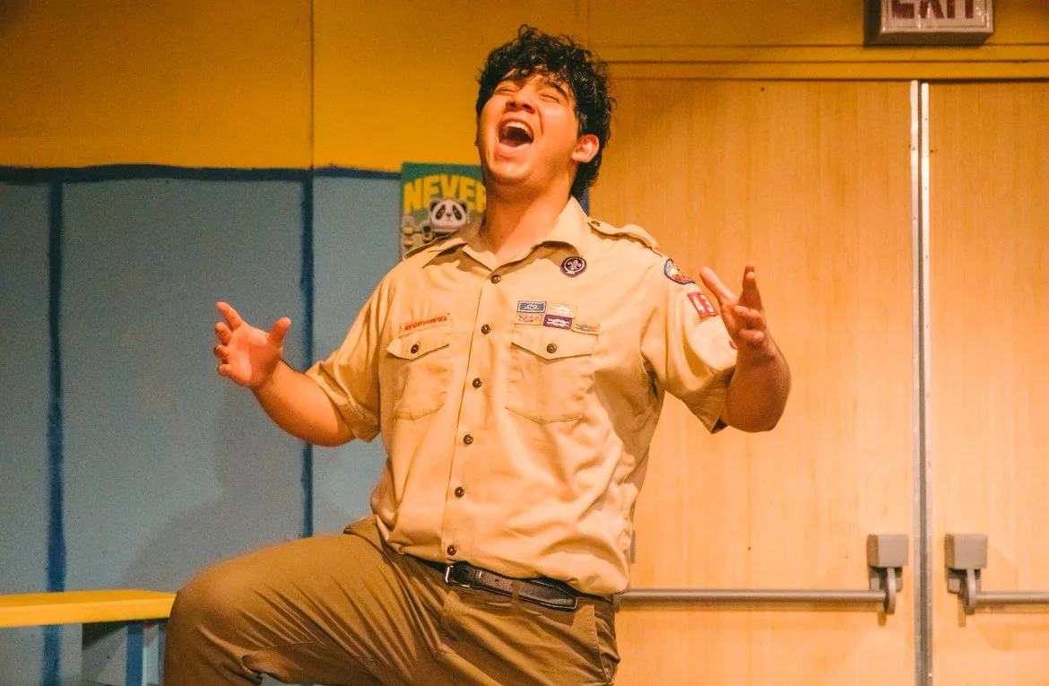 Actor in a boy scouts outfit singing on a colorful stage