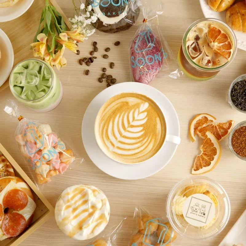 A flat lay of coffee, desserts, and snacks on a light wooden table. Includes a cup of latte with latte art, a glass of iced coffee with orange slices, assorted cookies, muffins, and small bowls of colorful toppings, with some flowers and scattered coffee beans.