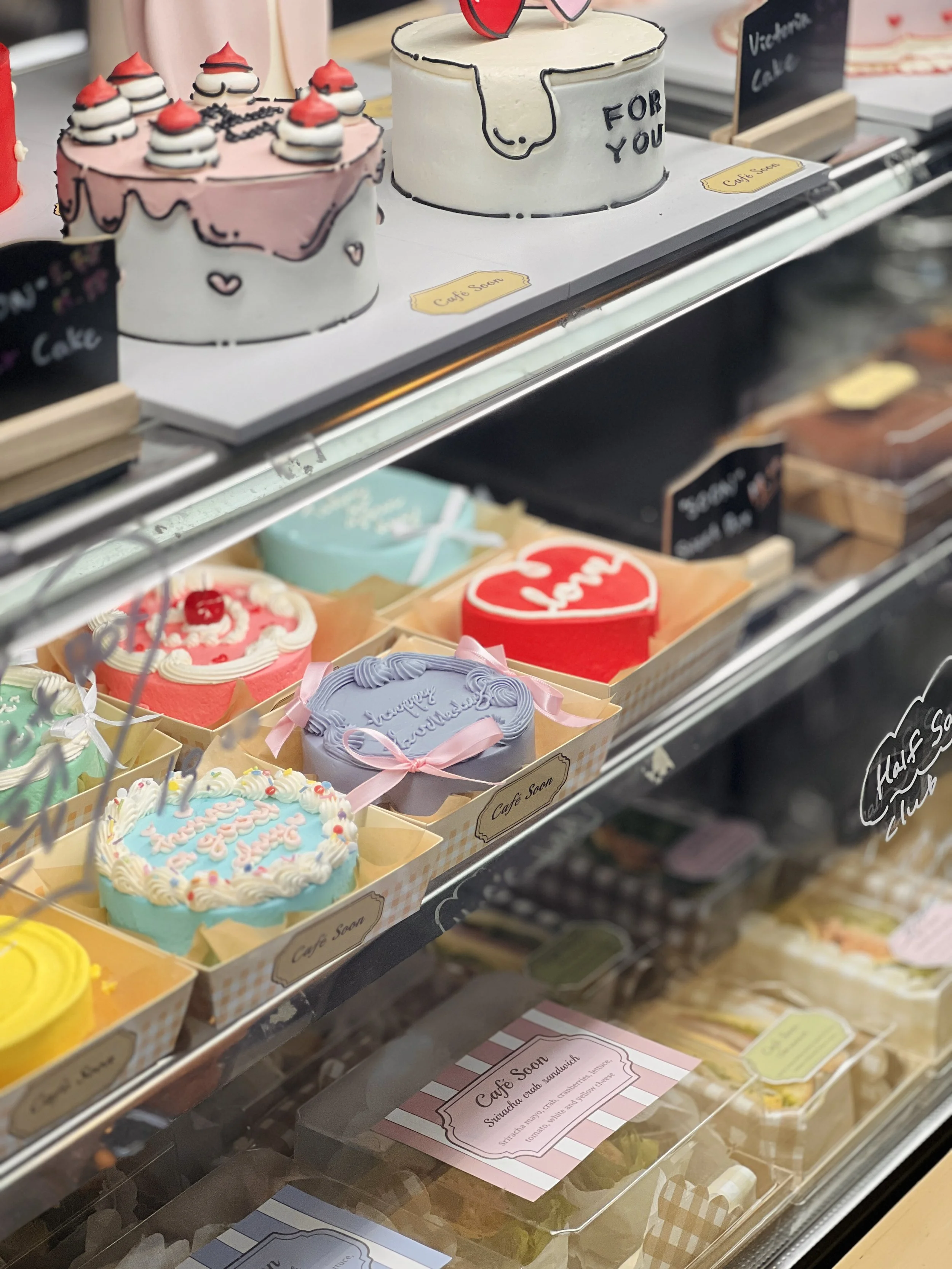 Display case with various colorful cakes, including a pink cake with whipped topping and cherries, a white cake with a dog design and glasses, and smaller decorated cakes with bows and messages