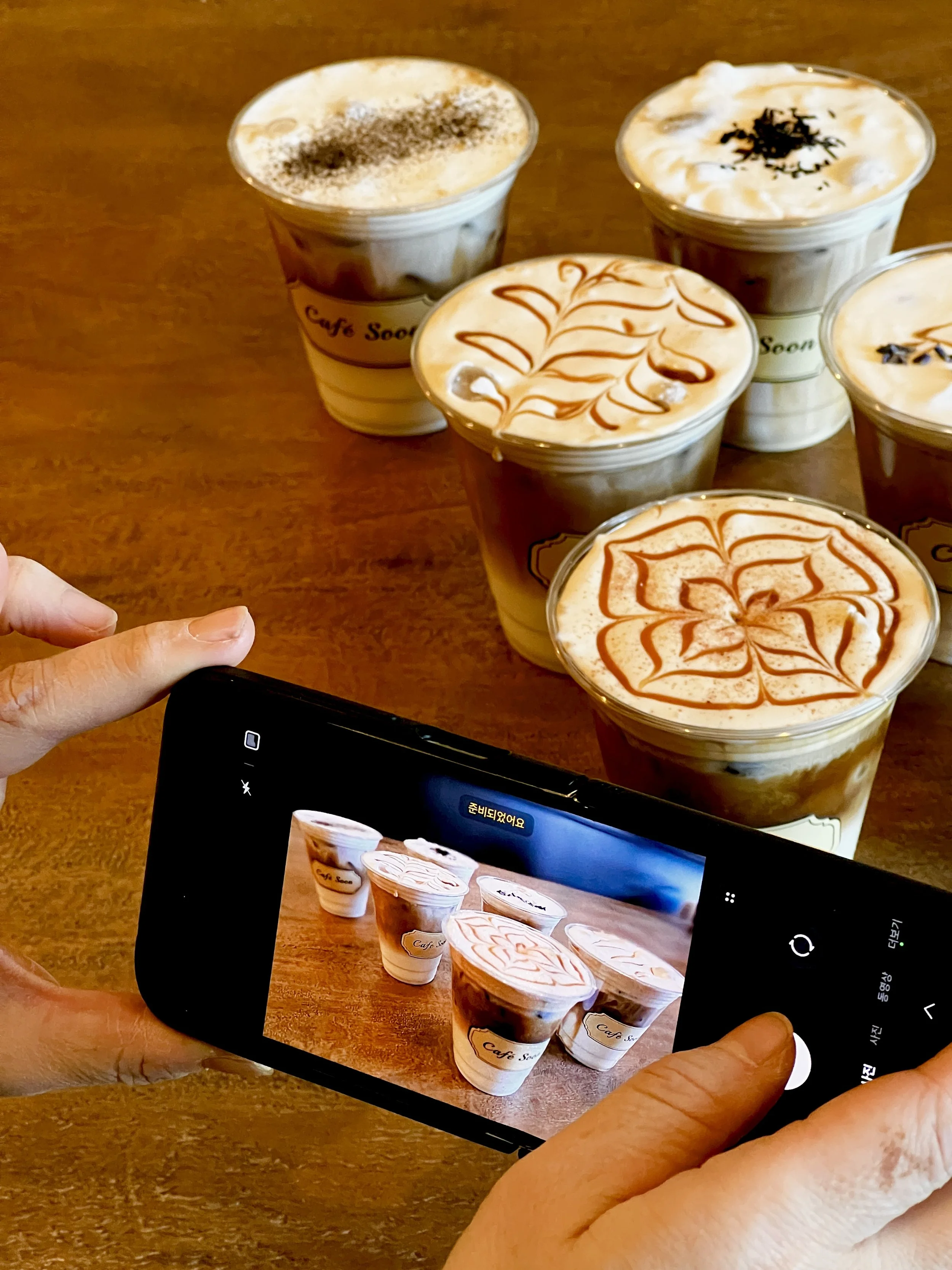 Person taking a photo of five different coffee drinks with a smartphone on a wooden table.