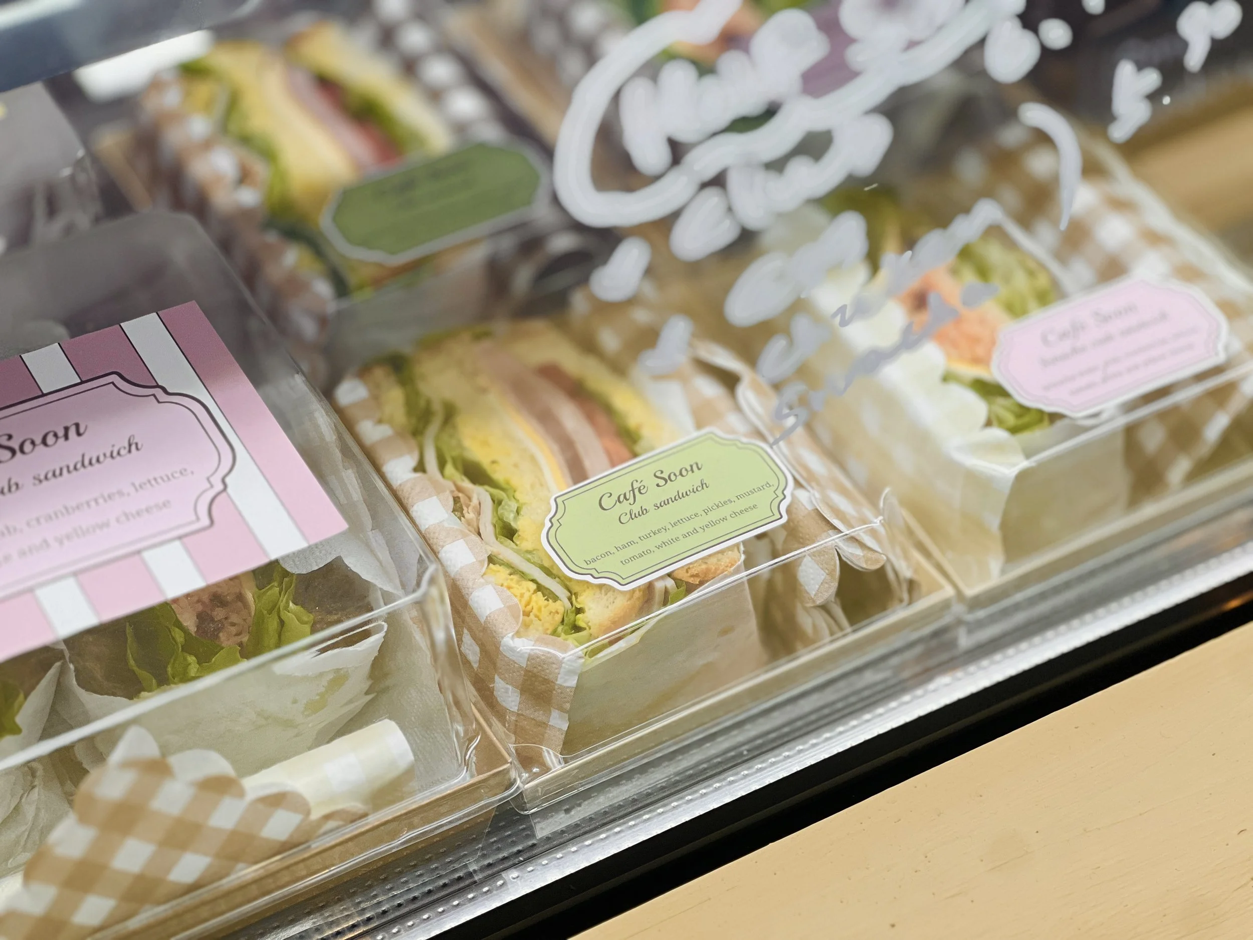 Assorted sandwich packs in a display case, including club sandwiches with various ingredients such as lettuce, turkey, and cheese, labeled 'Cafe Soon.'