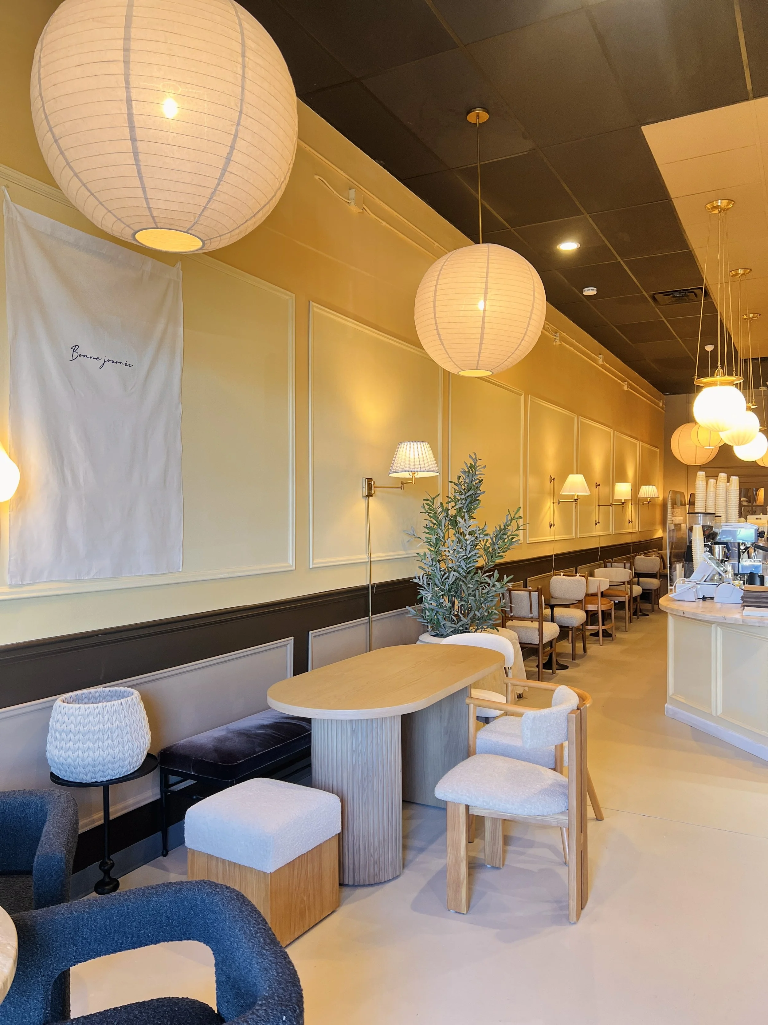 Interior of a cozy cafe with warm lighting, paper lantern hanging lamps, seating with various chairs and a small table, and a coffee counter in the background.