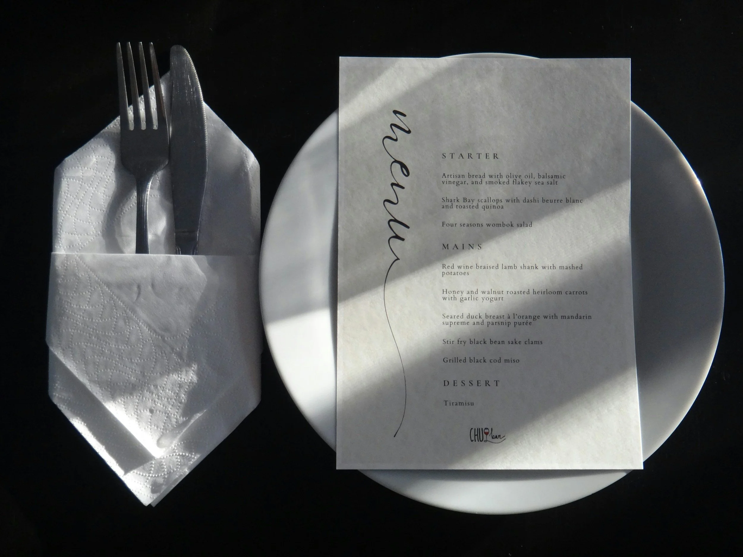 A white plate with a menu on top, a fork and knife wrapped in a napkin to the left, and a dark background.