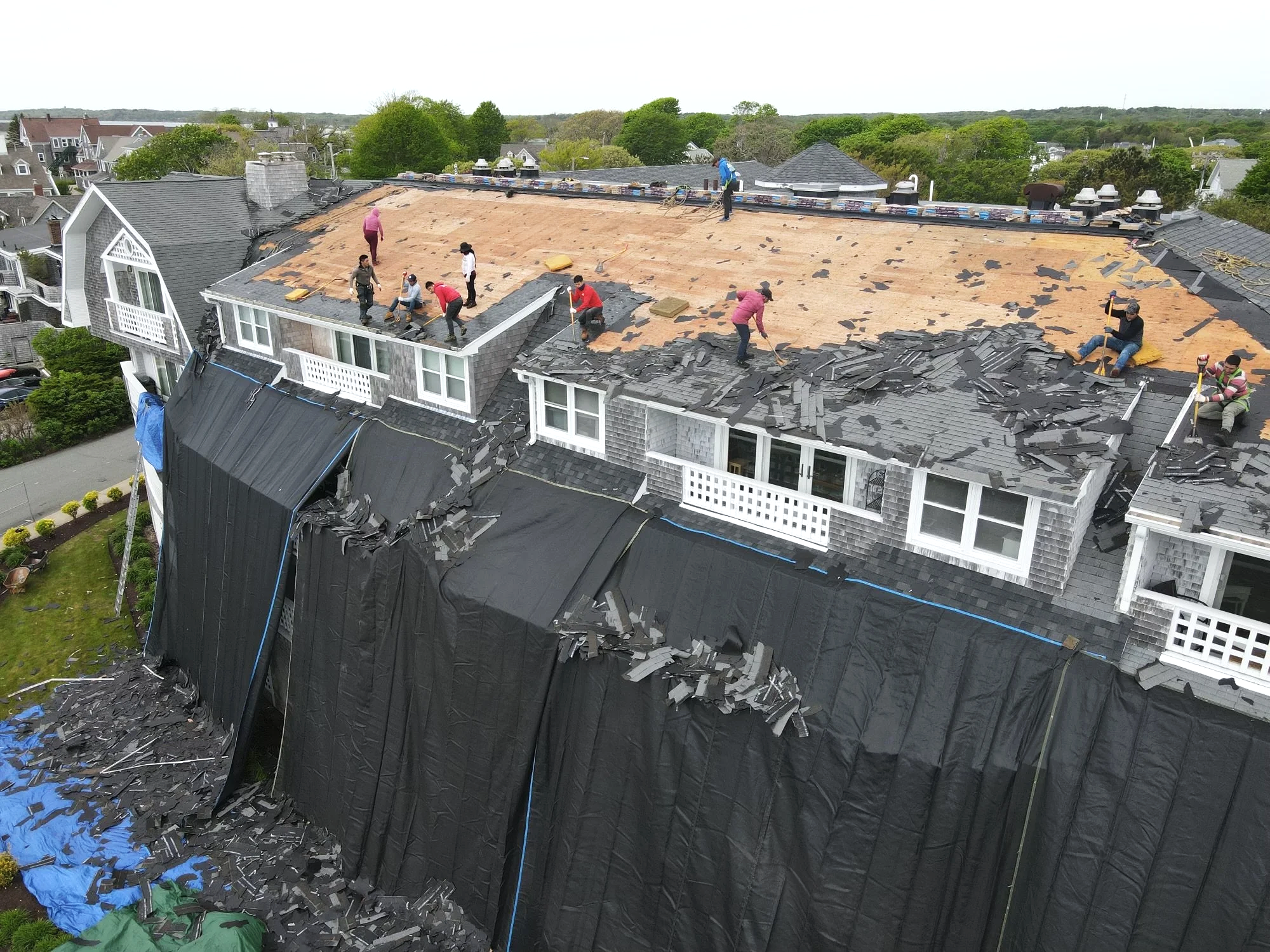 Construction workers on a roof removing shingles before replacing the roofing material, with parts of the old roof hanging down and debris on the ground.