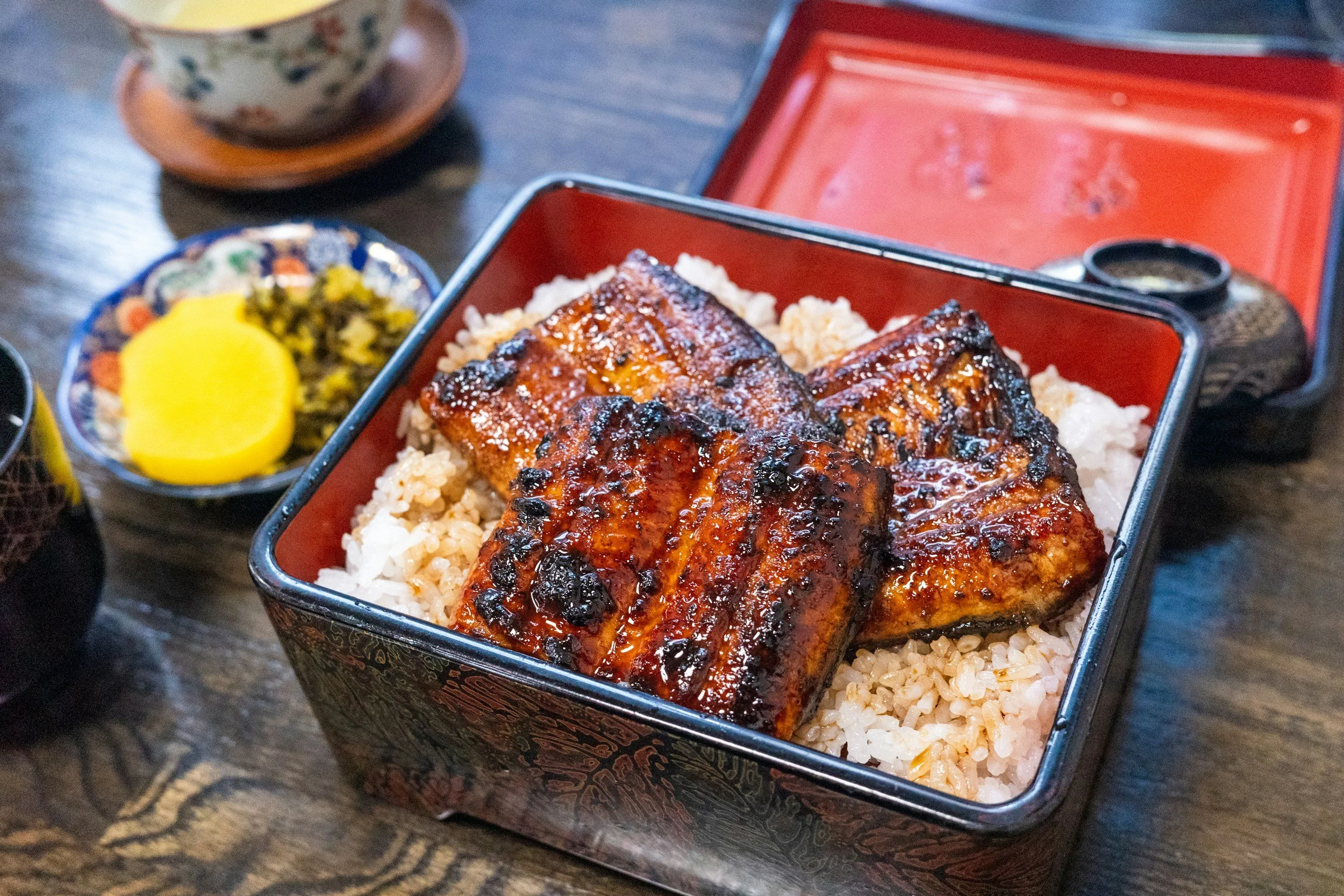 Bento box with grilled eel glazed with sauce on rice, with side dishes including yellow pickled radish and greens, on a wooden table.