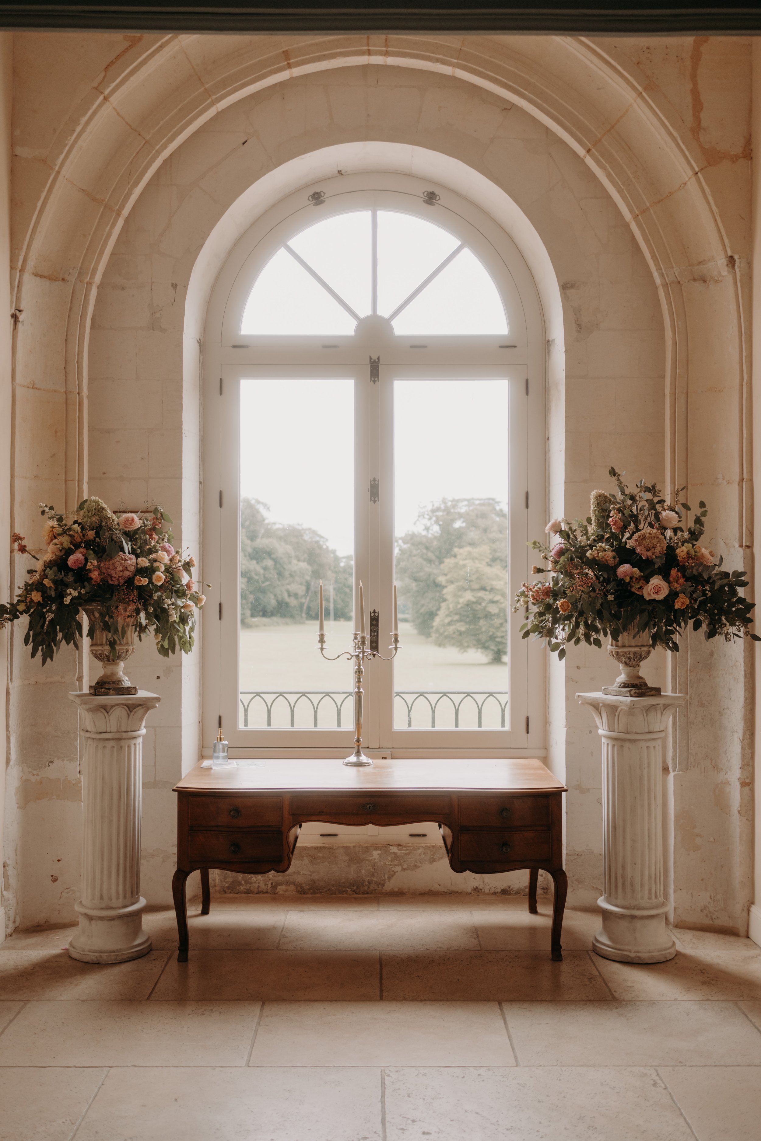 Une grande fenêtre avec un cadre blanc, entourée de murs en pierre, avec deux grands vases de fleurs sur des socles classiques de chaque côté, une table en bois au centre avec un chandelier.