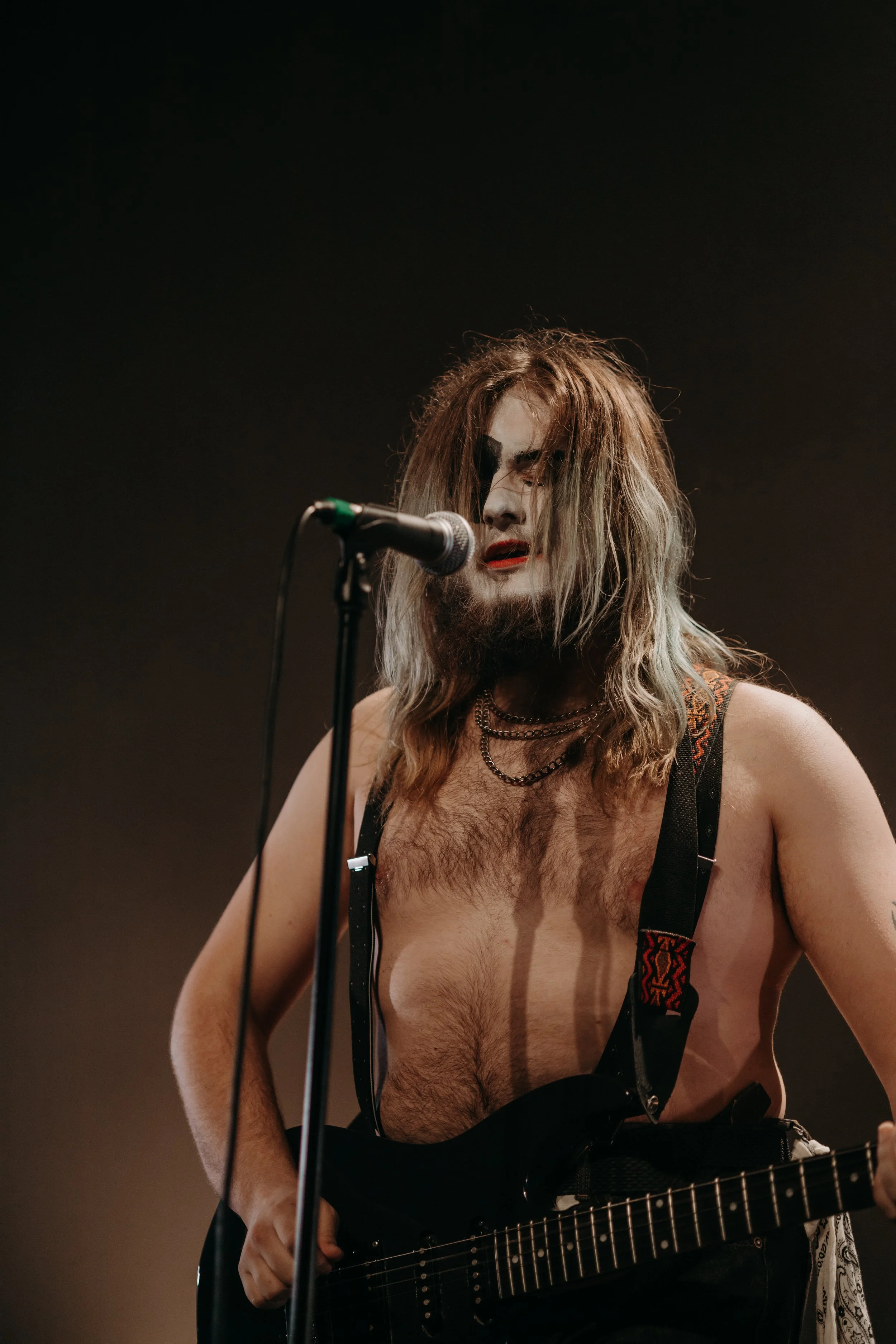 Un homme avec des cheveux longs et un maquillage sombre joue de la guitare et chante dans un micro, vêtement sans chemise, avec des suspenders et plusieurs colliers, dans un environnement sombre et à l'éclairage tamisé.