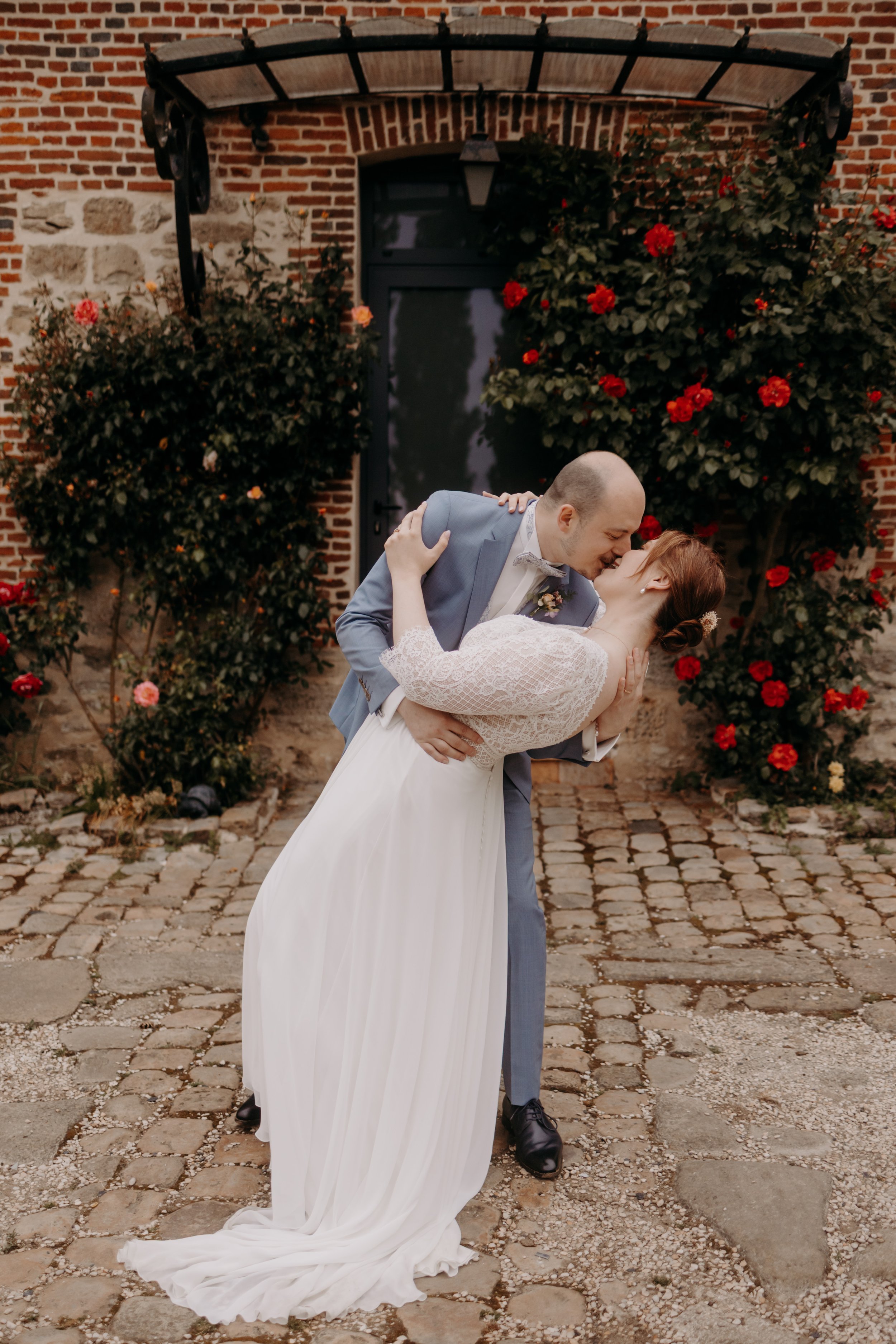 Un couple marié s'embrassant lors de leur mariage, avec un décor de fleurs rouges et un mur en briques en arrière-plan.