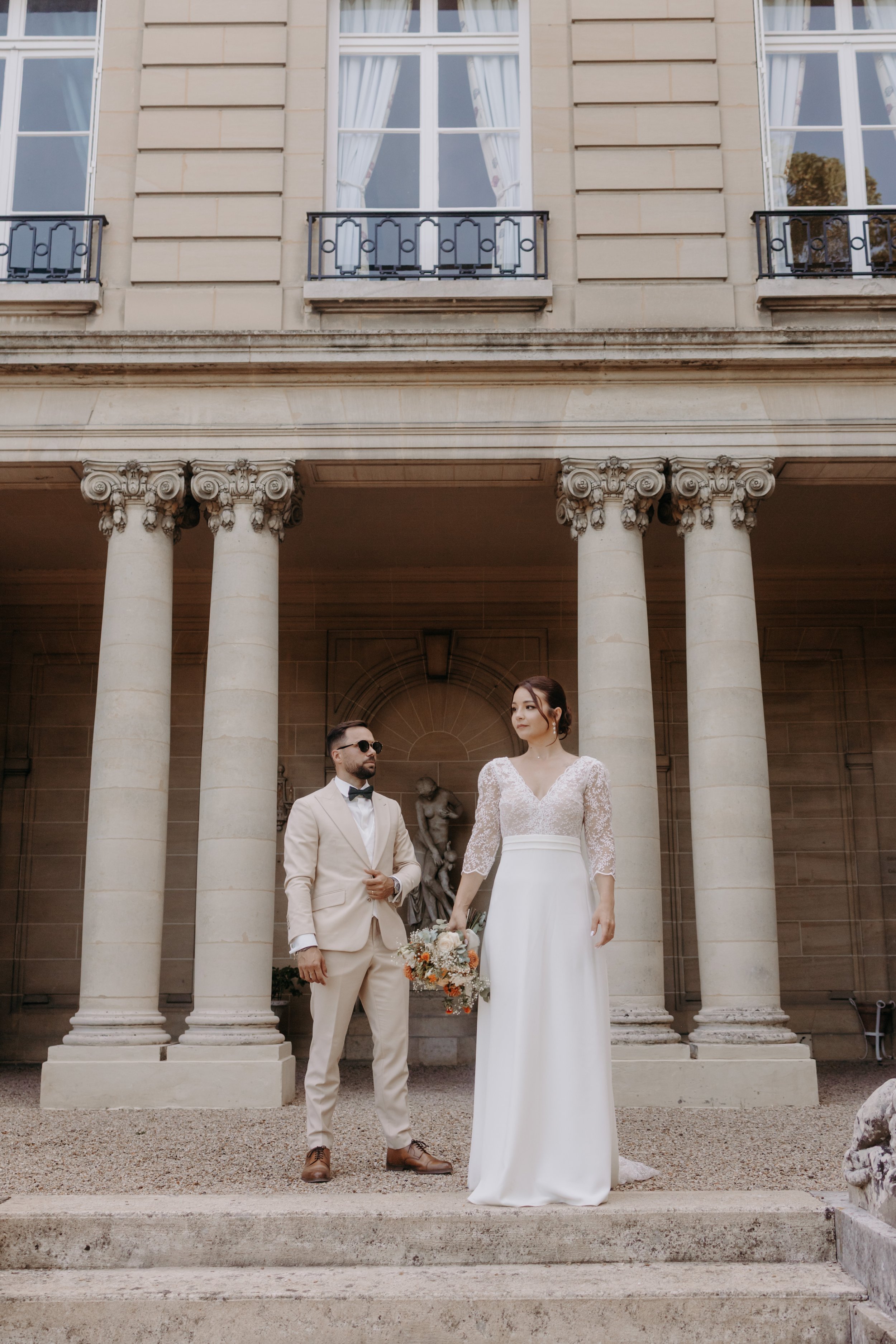 Un couple en mariage posant devant un bâtiment en pierre avec des colonnes, la femme porte une robe blanche avec un bouquet, l'homme en costume beige avec un nœud papillon.