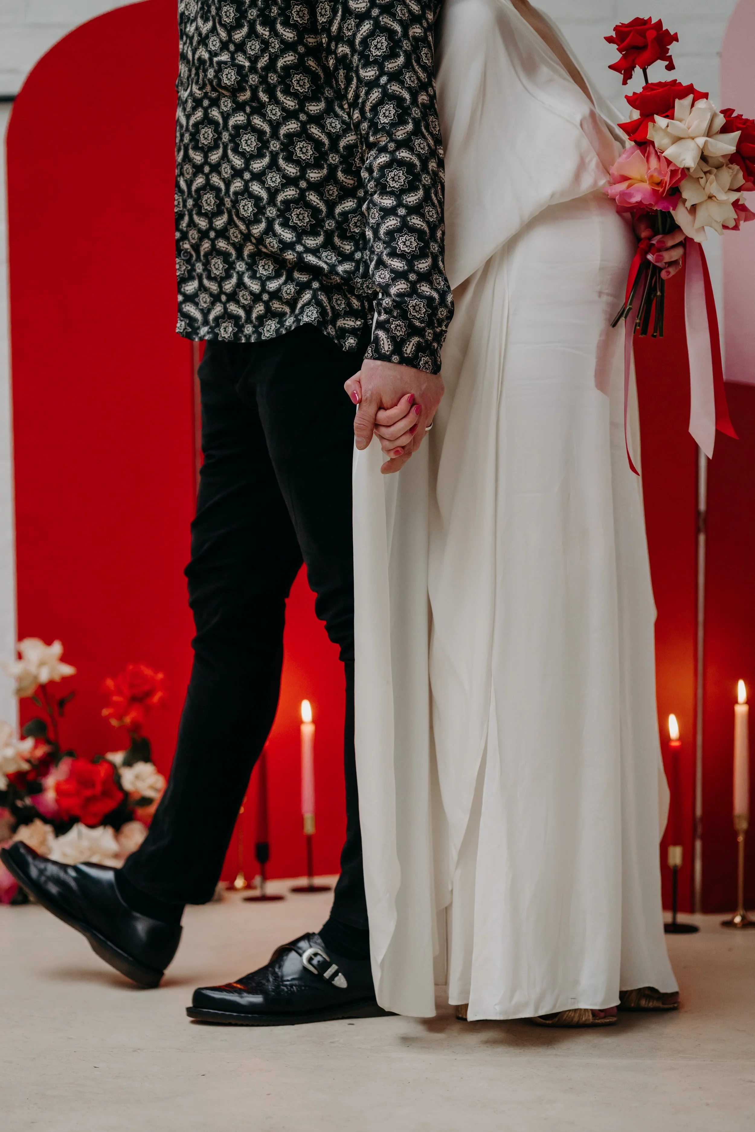 Un couple marié se tenant la main lors de leur mariage, la femme portant une robe blanche et tenant un bouquet de fleurs, avec des bougies allumées en arrière-plan.