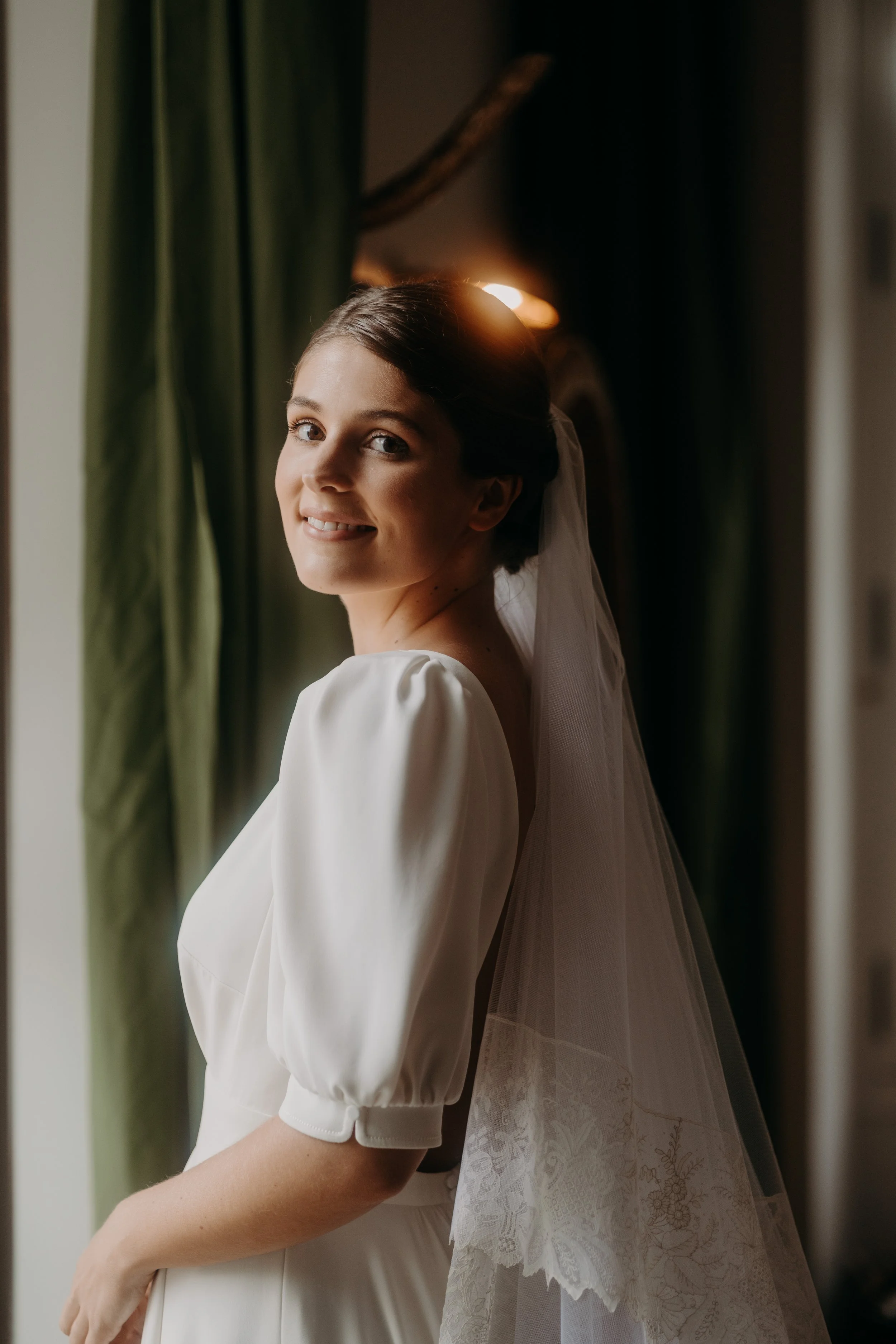 Jeune femme souriante en robe blanche et voile de mariée, debout près d'une fenêtre, fond vert et décor ancien.