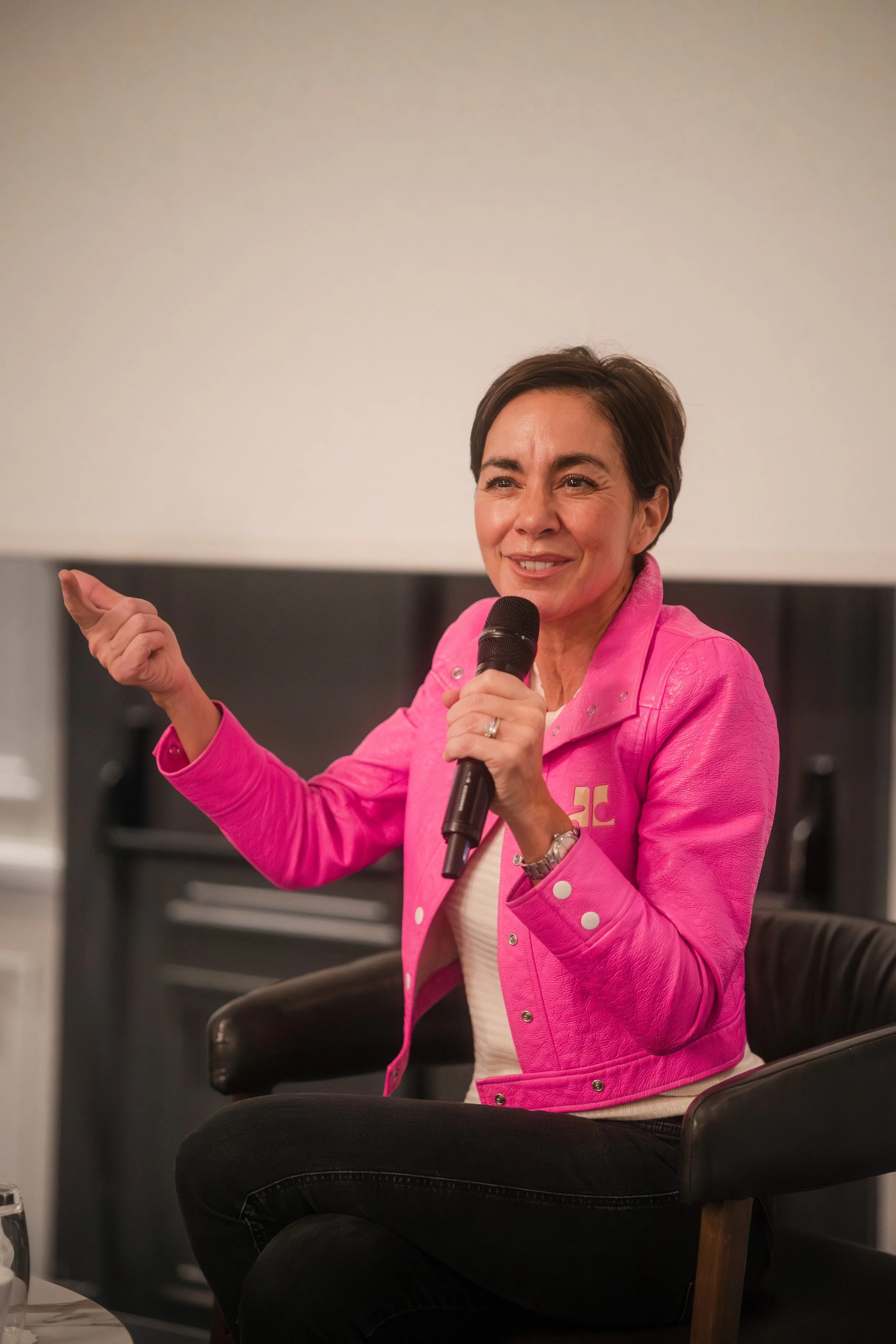 Une femme avec une veste rose, assise dans un fauteuil, parle en tenant un microphone, lors d'une discussion ou d'une interview.