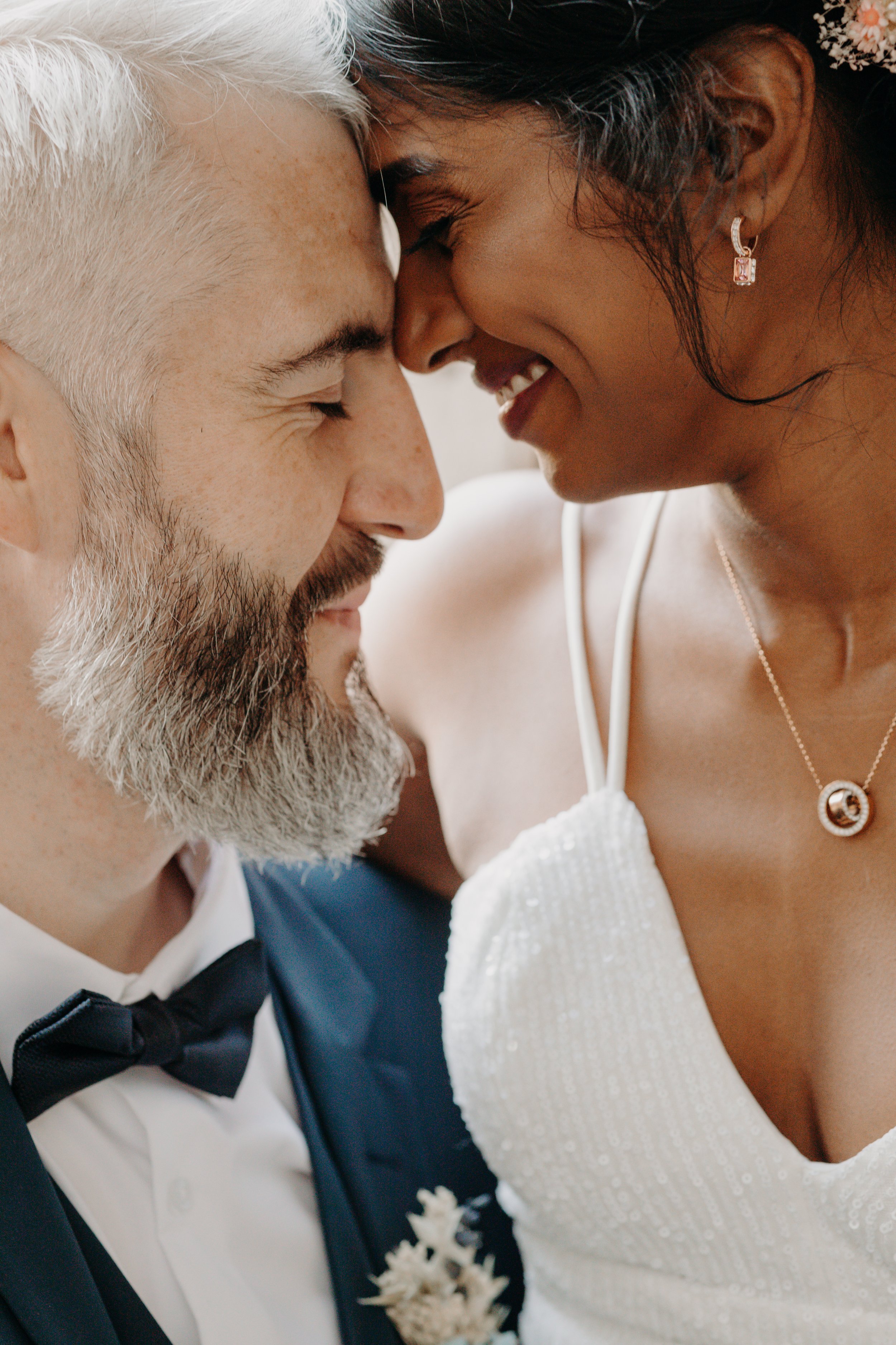 Un couple en tenue de mariage se touche le front, souriant tendrement, lors d'une coiffure ou d'une séance photo de mariage.