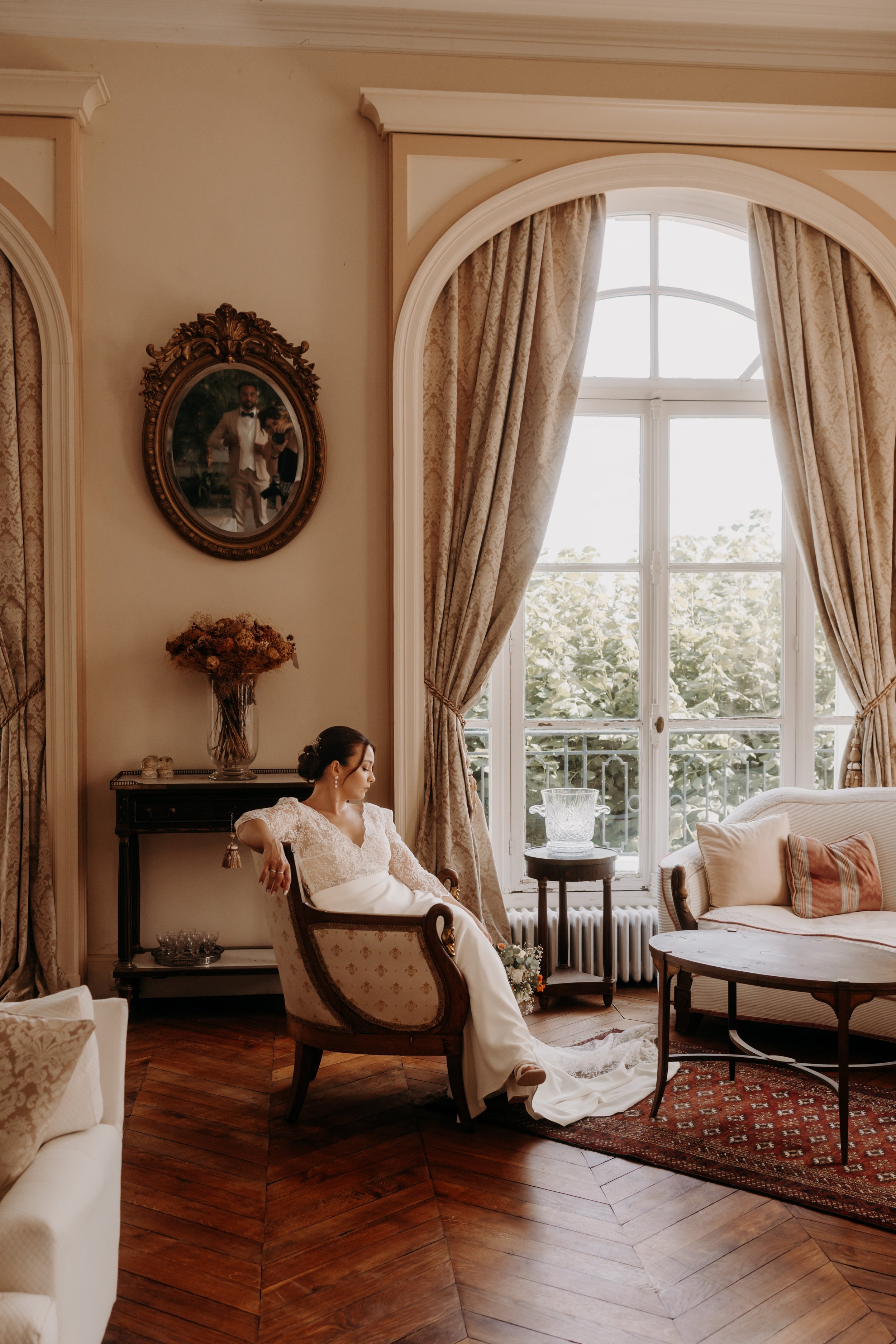 Une femme en robe de mariée assise dans un salon élégamment décoré avec de grandes fenêtres, des rideaux épais et des meubles vintage.