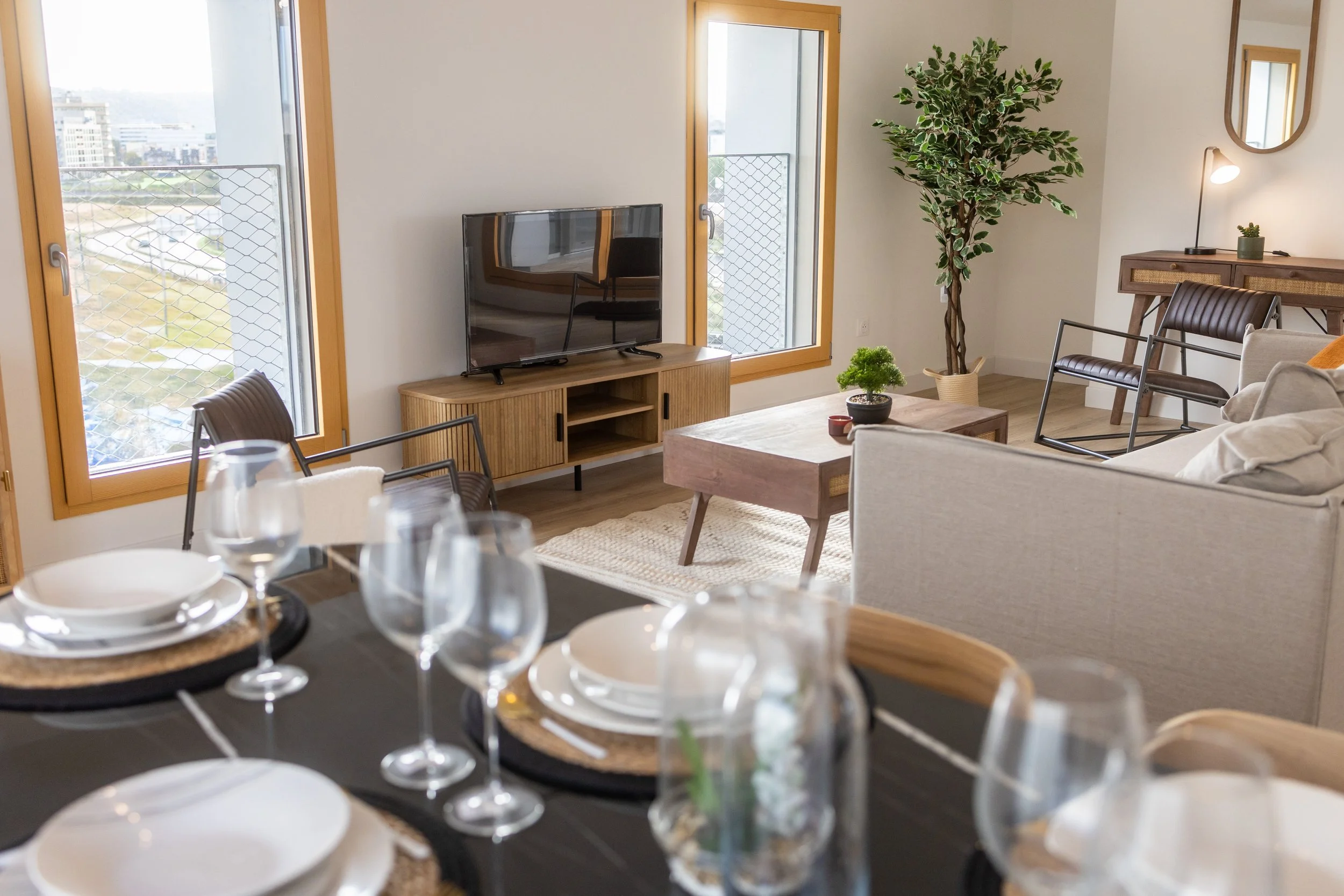 Salon de salon avec canapé, fauteuils, table basse, grande plante, télévision, et fenêtres avec vue urbaine