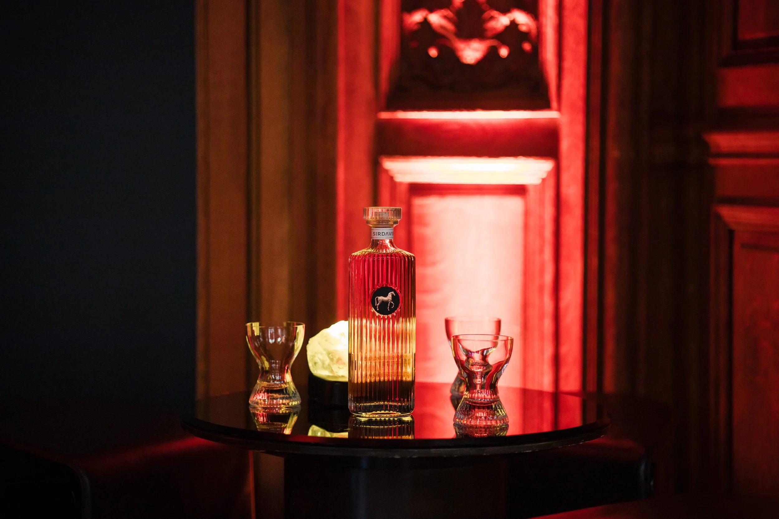 Une bouteille de parfum rouge avec un logo de cheval noir, entourée de trois petits verres et un objet blanc lumineux, sur une table noire, arrière-plan en bois avec éclairage rouge