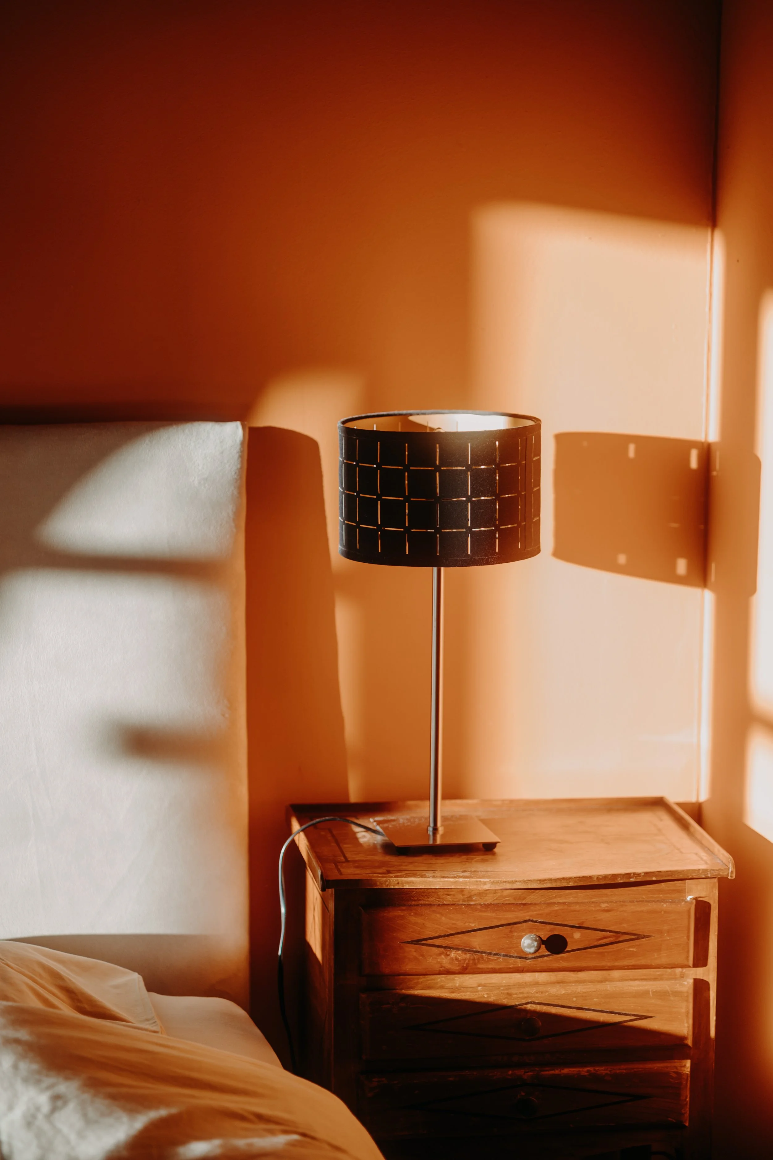Lampe de chevet sur une table en bois dans une chambre, éclairant par la lumière douce et créant des ombres sur le mur.