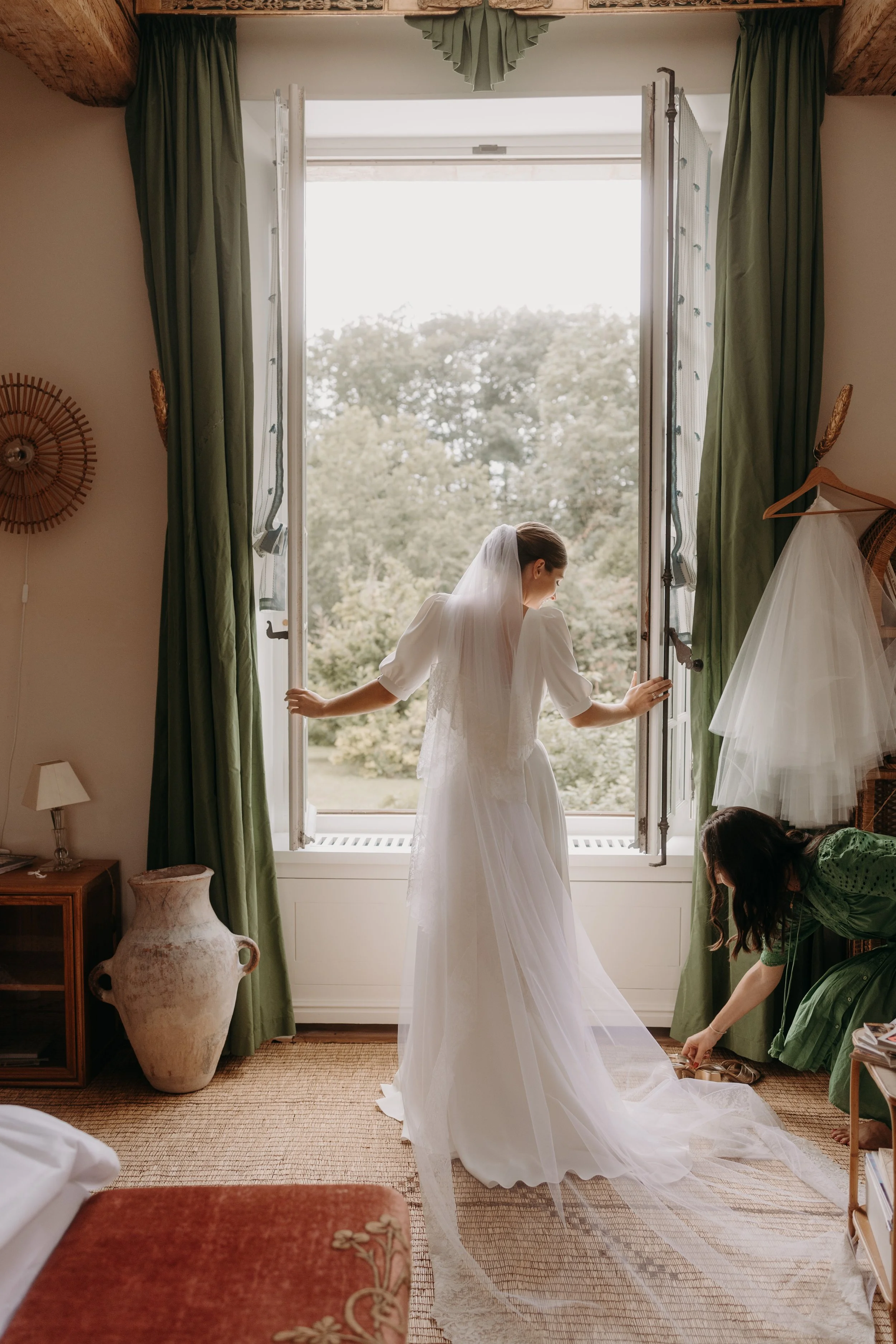 Une mariée en robe blanche se tient près d'une fenêtre ouverte, une femme ajustant ses chaussures à ses pieds, dans une chambre décorée avec des rideaux verts et des meubles en bois.
