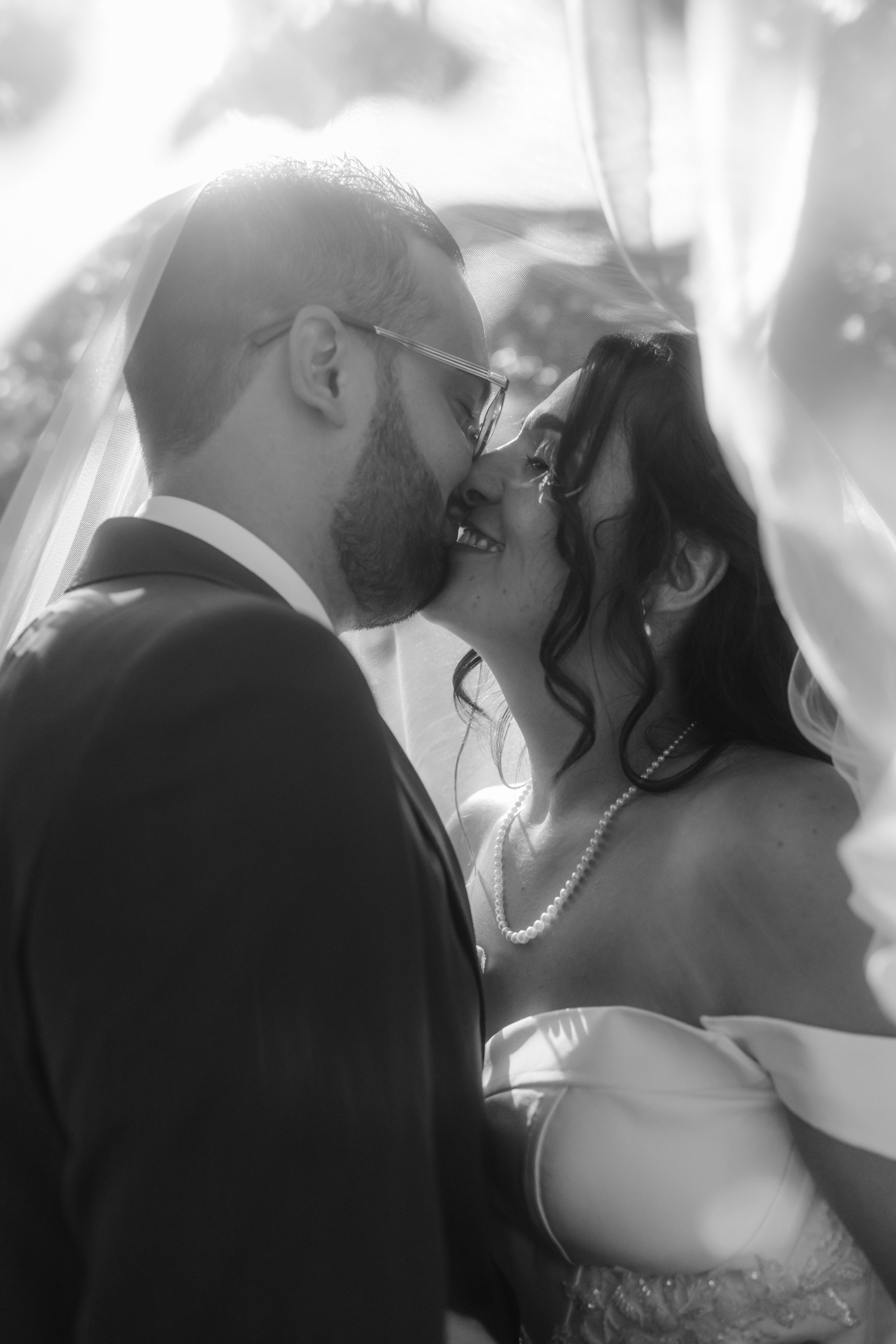 Un couple de mariés s'embrasse sous un voile de mariée, en noir et blanc.