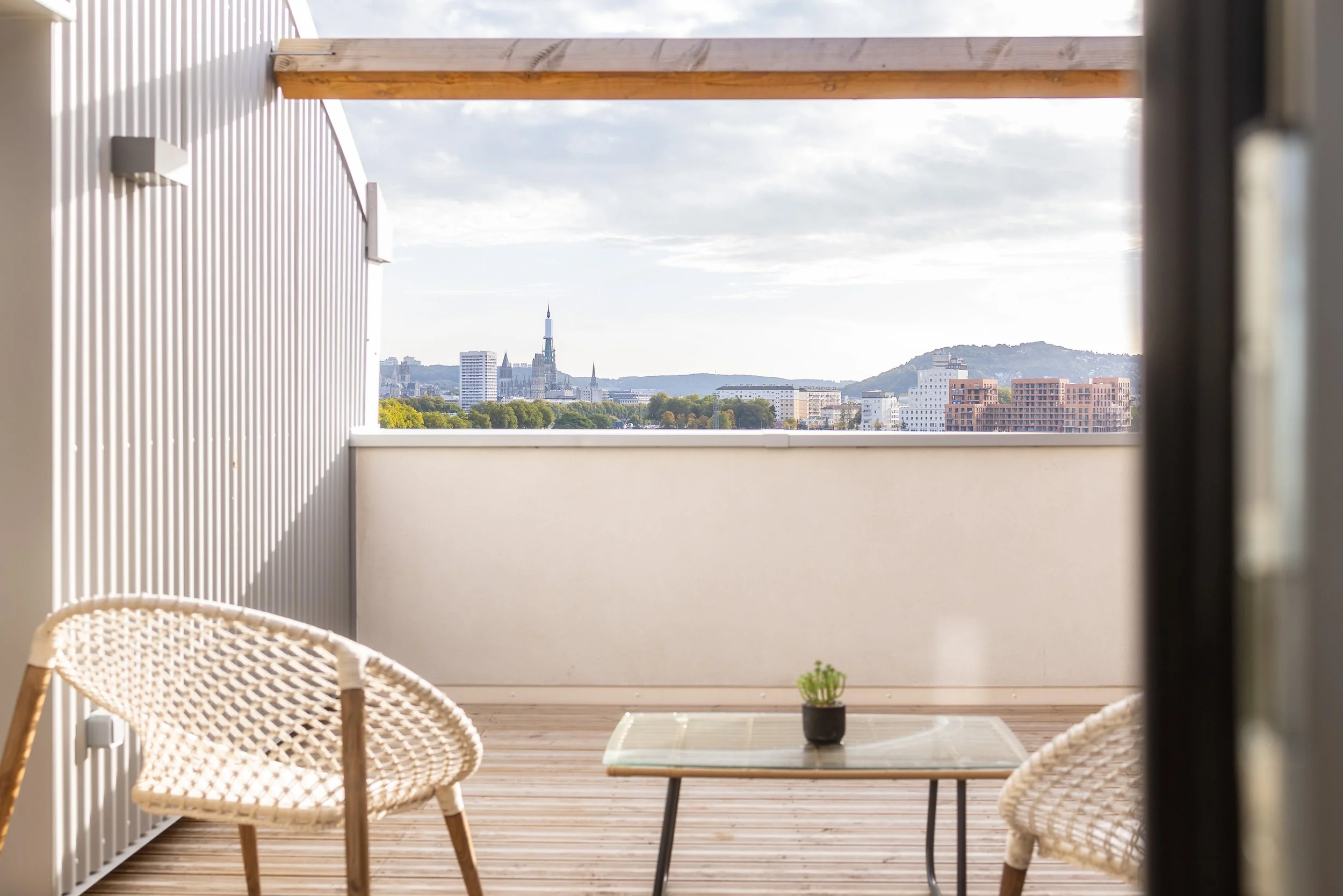 Vue depuis un balcon avec deux chaises en osier, une table en verre et un petit pot de plantes, avec une ville en arrière-plan.