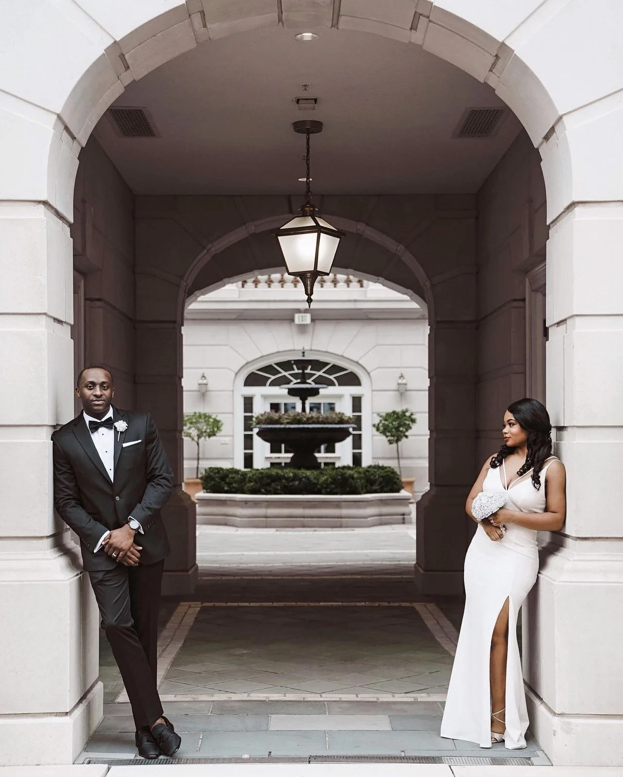 A man in a black tuxedo and a woman in a white evening gown holding a bouquet, standing under an archway in front of a fountain at a formal event.