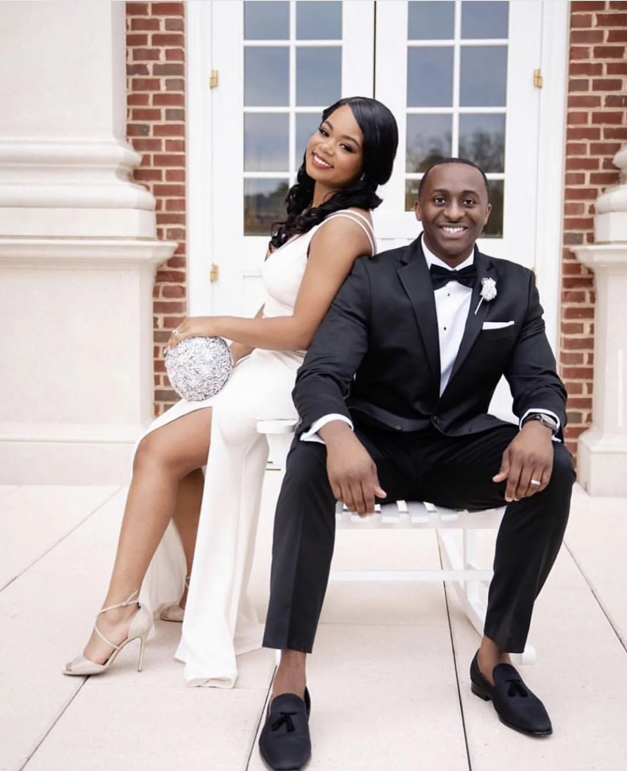 A couple in formal attire, the woman in a white dress holding a glittery clutch, and the man in a black tuxedo, sitting on a white bench outside a building with brick and stone columns, standing in front of a glass door.