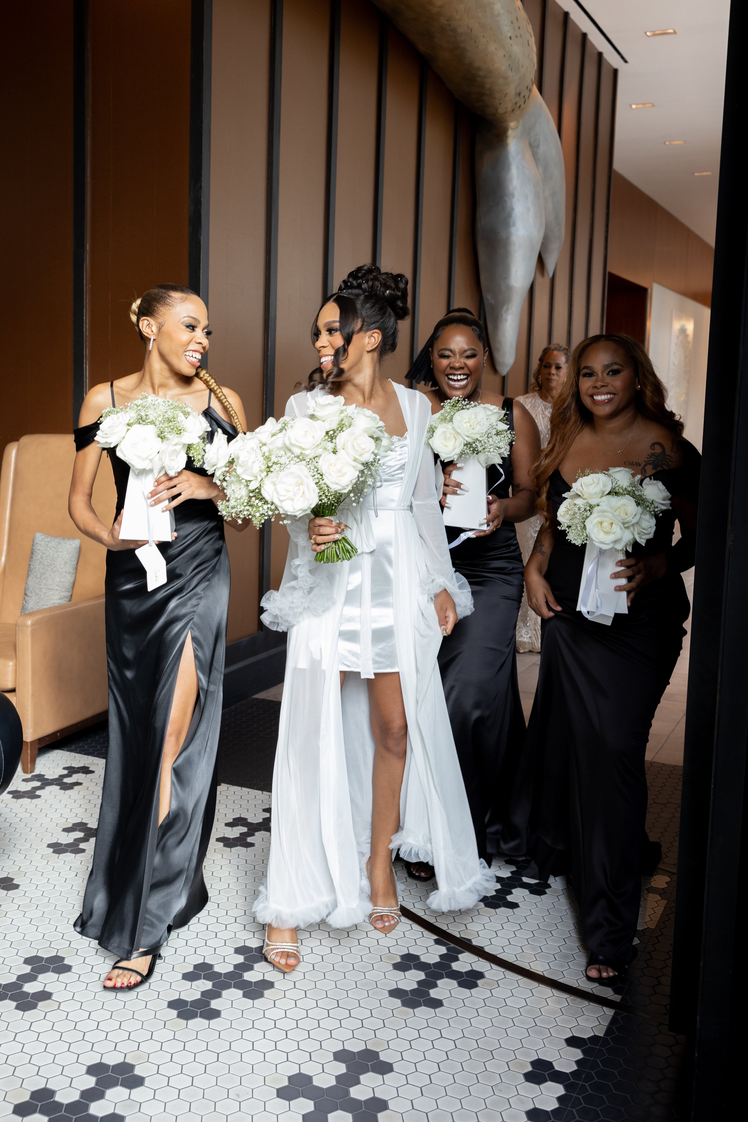 A bride and her bridesmaids smiling and holding bouquets of white flowers in a hotel lobby.